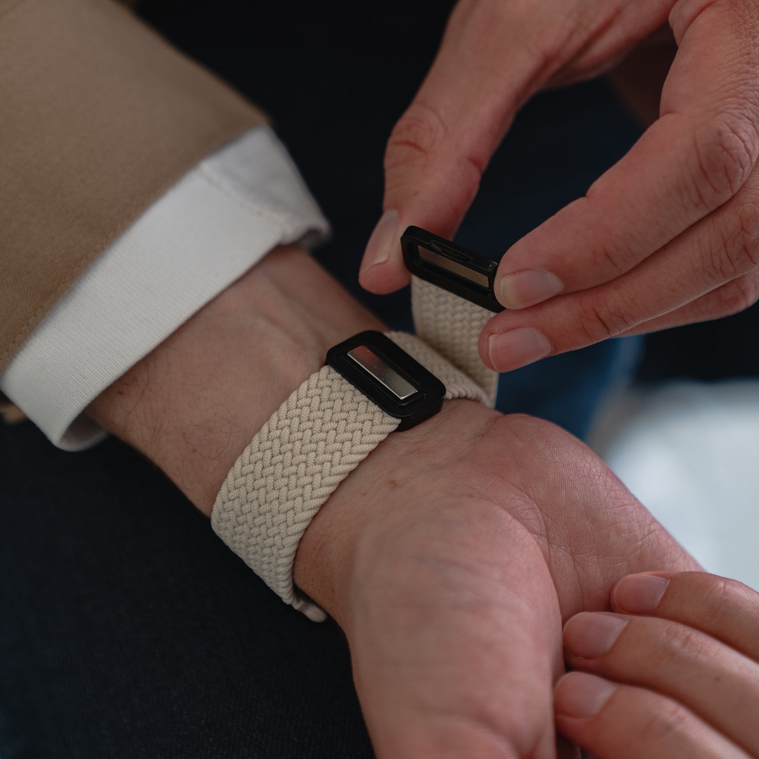 Eine Person befestigt das arktisband Geflochtene Apple Watch Armband „Casual“ mit Magnetverschluss in Beige und Schwarz um ihr linkes Handgelenk, kombiniert mit einer hellbraunen Jacke und einem weißen Hemd.