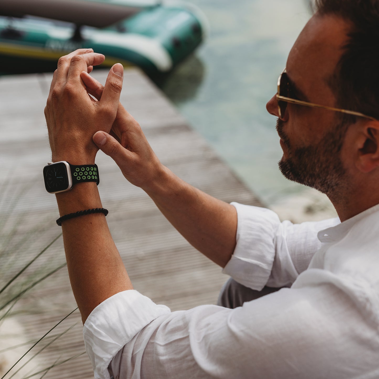 Ein Mann mit weißem Hemd, Sonnenbrille und einem Arktisband Apple Watch Silikonarmband „Energy“ in Schwarz und Grün sitzt draußen am Wasser und blickt auf seine Hände, hinter ihm ein Holzsteg und ein Schlauchboot.