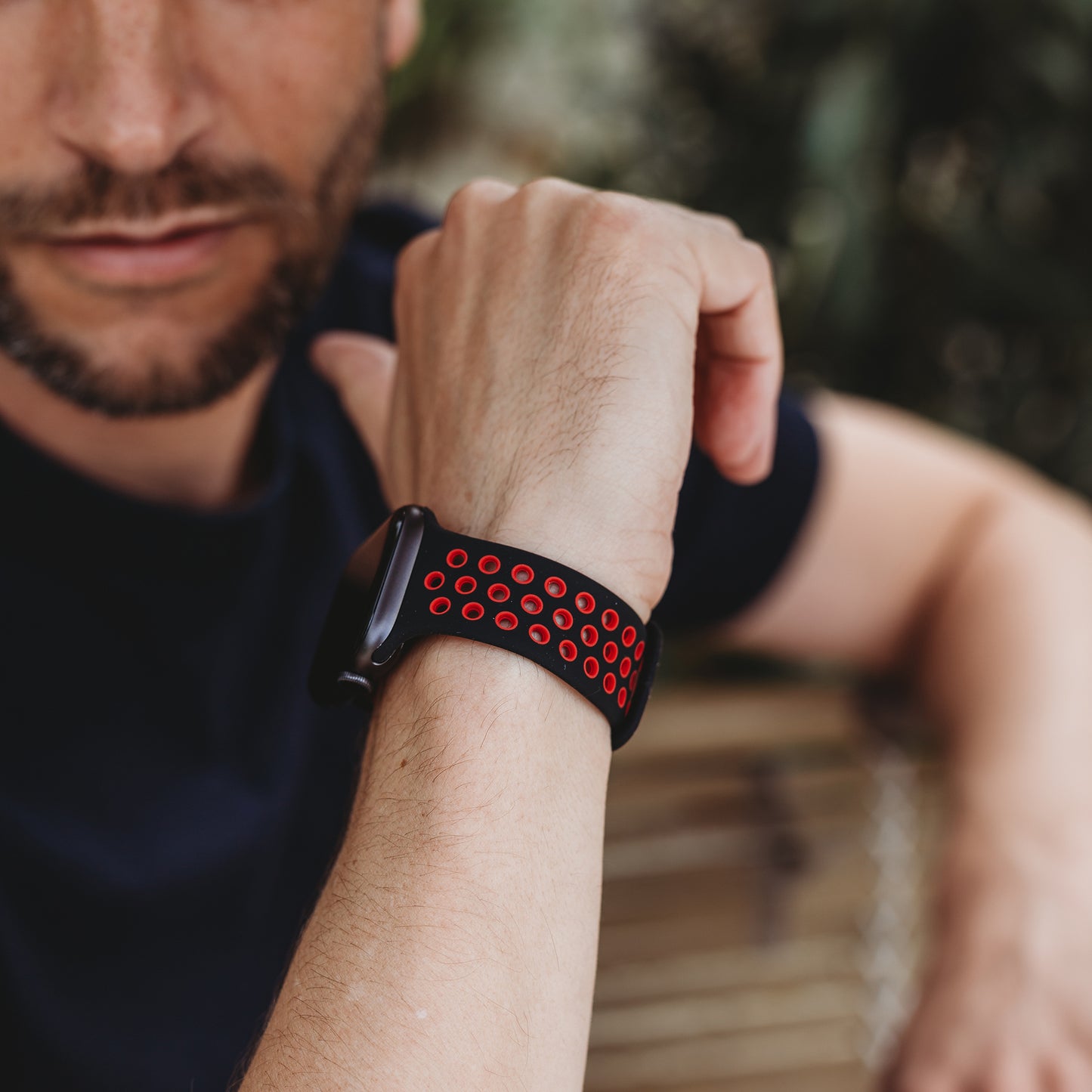 Eine bärtige Person in einem marineblauen Hemd ruht mit dem Arm und trägt das arktisband Apple Watch Silikonarmband "Energy" von arktisband in Schwarz und Rot. Der Hintergrund ist unscharf.