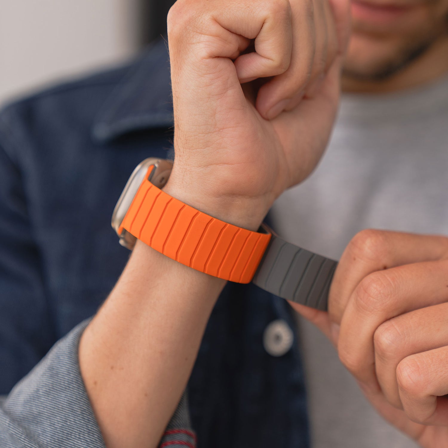 Eine Person in Jeansjacke und grauem Hemd rückt ein magnetisches Arktisband Apple Watch MagFlow-Armband mit flexiblem Silikon und zweifarbigem orange-grauem Design an ihrem Handgelenk zurecht.