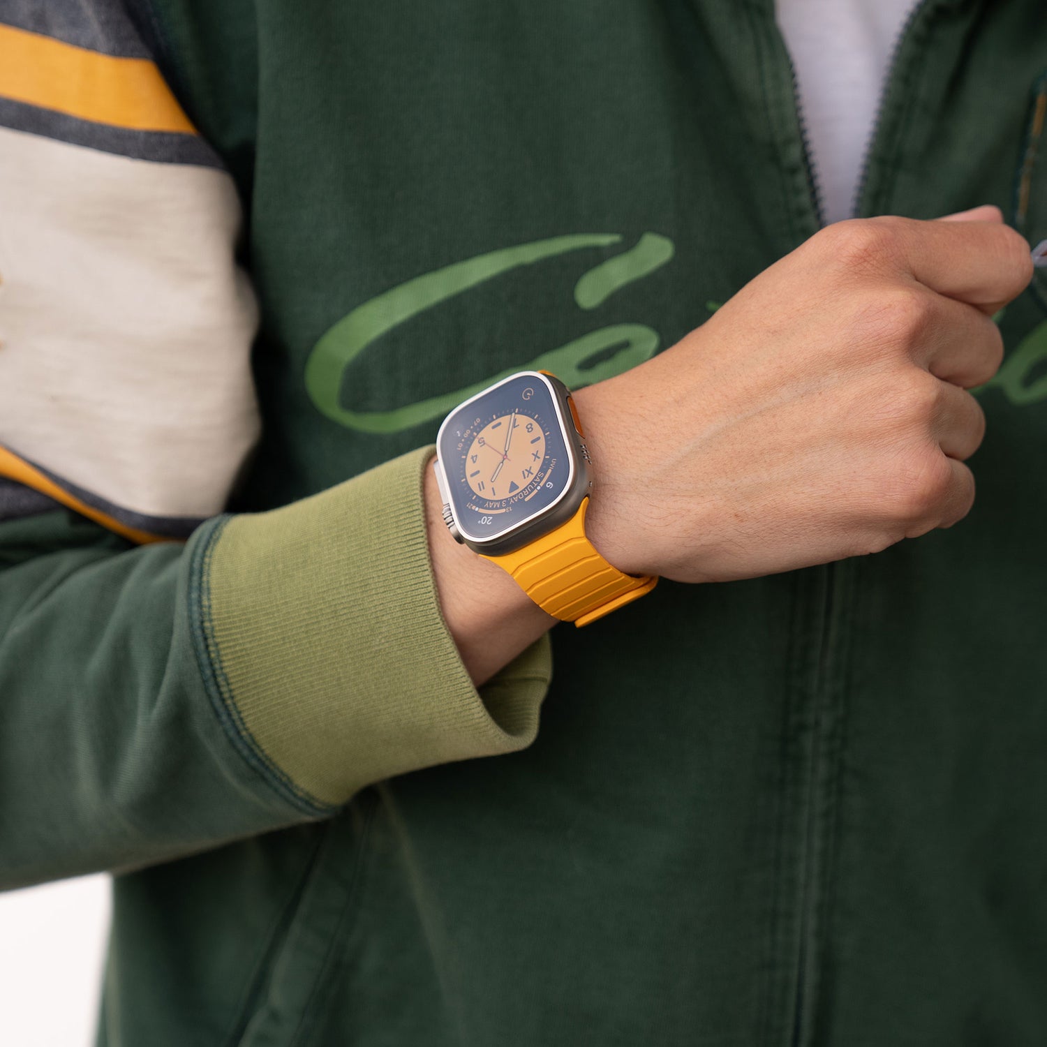 Eine Person in einer grünen Jacke ist von den Schultern abwärts zu sehen, die linke Faust ist geballt und erhoben, und sie trägt ein magnetisches Armband „MagFlow“ der Apple Watch mit einem gelben Band am Handgelenk.