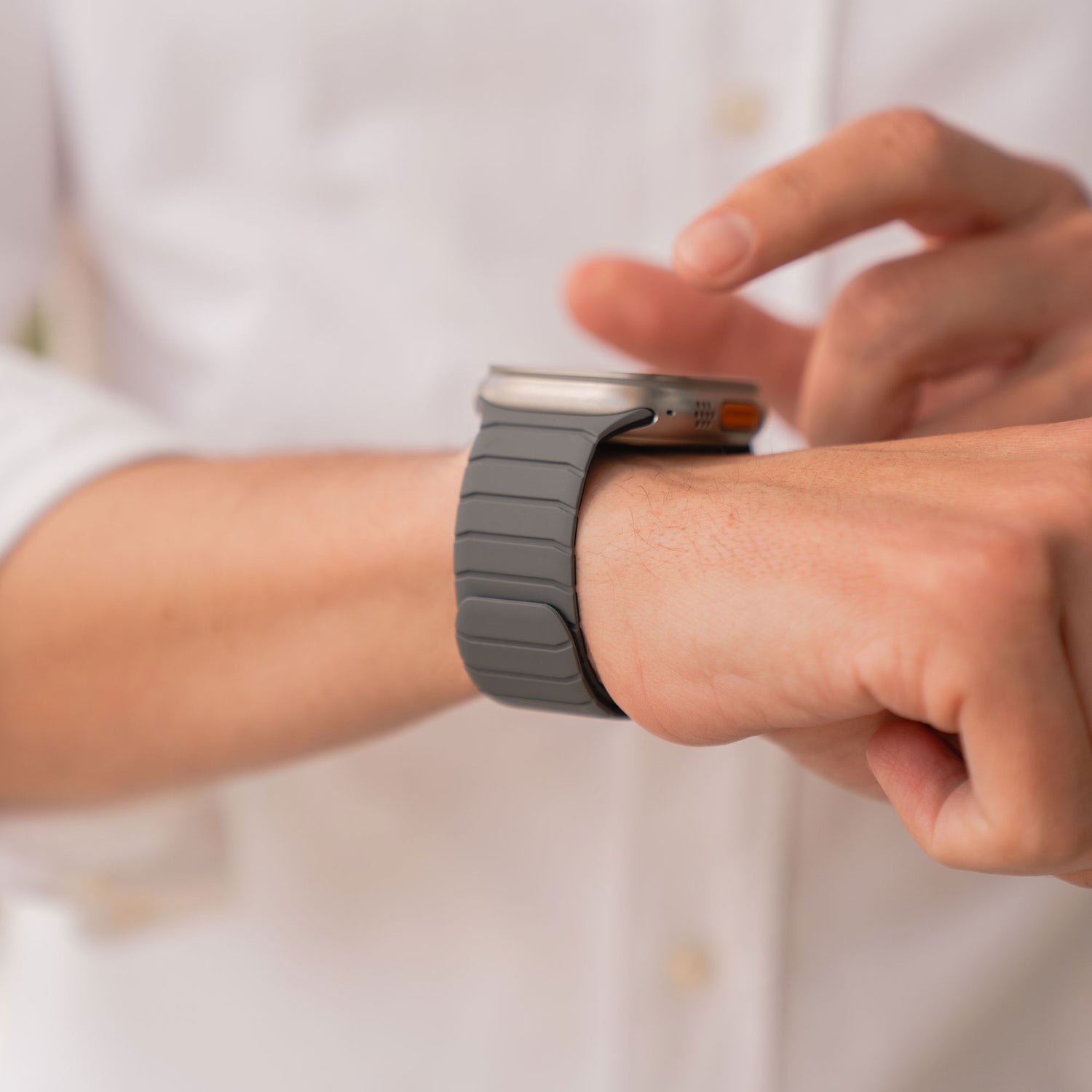 Eine Person im weißen Hemd tippt auf ein magnetisches Armband „MagFlow“ der Apple Watch mit grauem, geschmeidigem Silikonband, das sie am linken Handgelenk trägt.