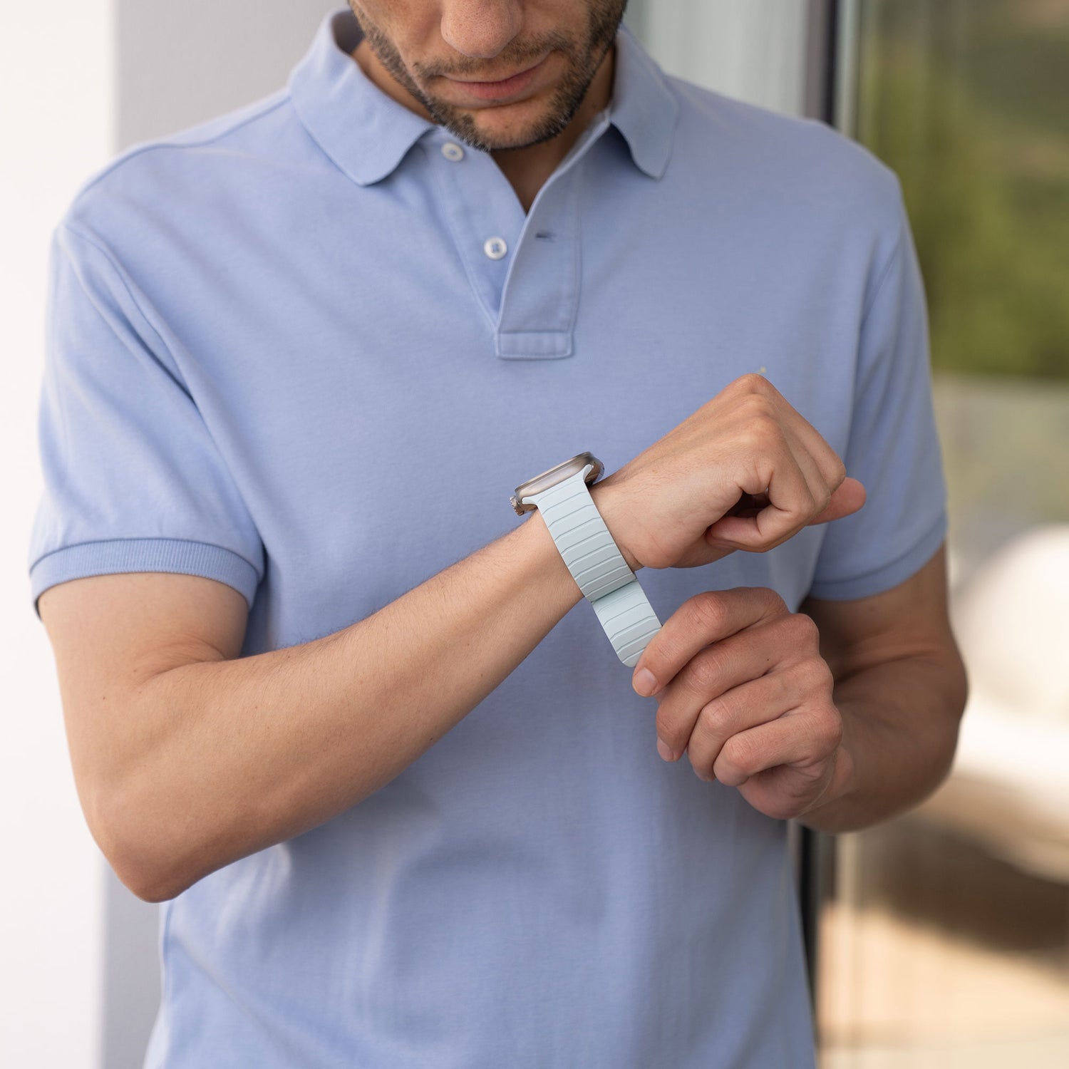 Eine Person in einem hellblauen Poloshirt passt ein magnetisches Apple Watch-Armband „MagFlow“ an ihrem linken Handgelenk an, während sie in einem Innenbereich neben einer Glastür steht.