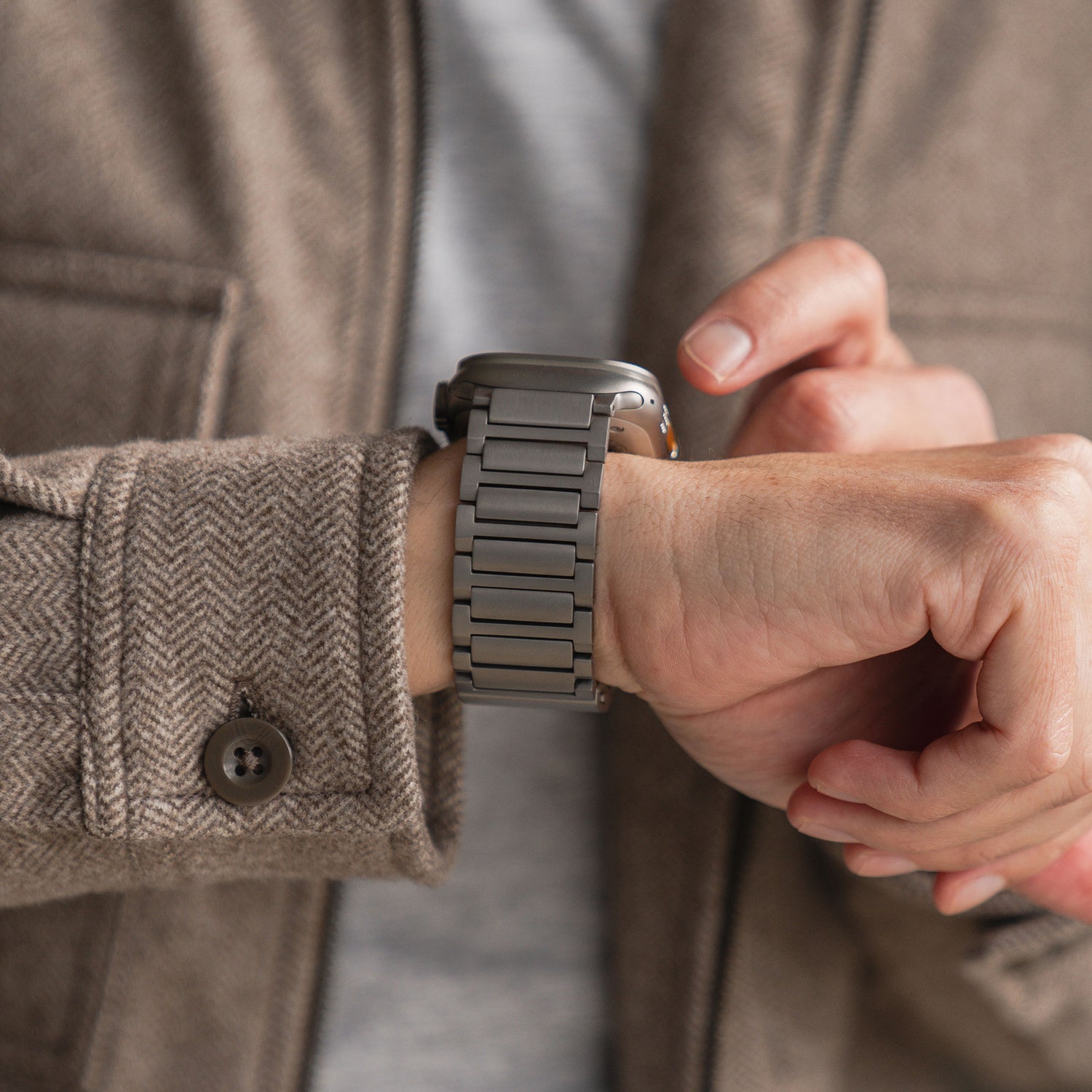 Eine Person in einer braunen Jacke mit Fischgrätenmuster checkt die Uhrzeit an ihrem linken Handgelenk. Sie trägt ein Arktisband Apple Watch Ultra Titanarmband „Maverick“. Auf dem Foto sind die Uhr und die Zeiger zu sehen.