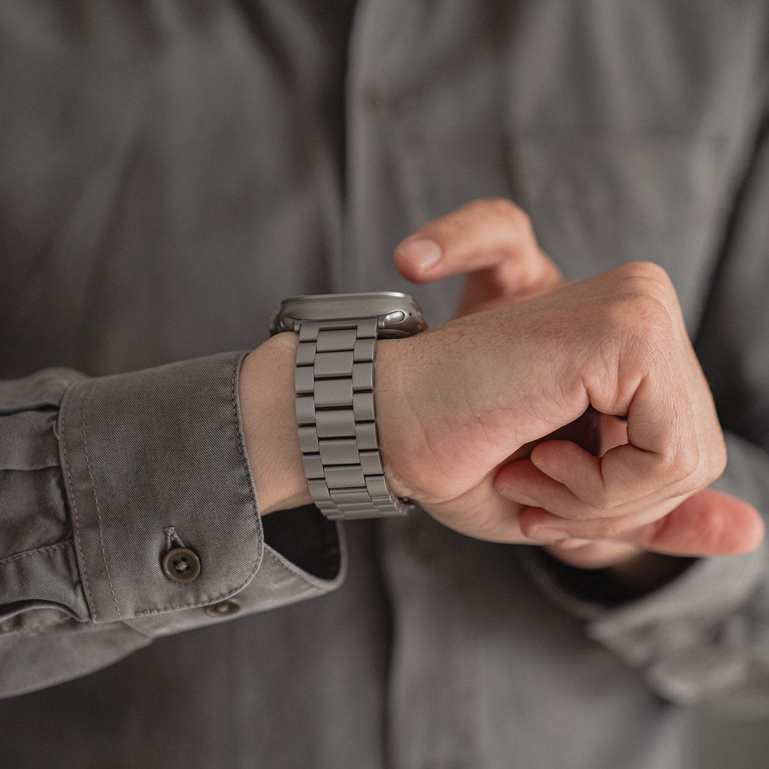 Eine Person in einem grauen Hemd mit Knöpfen berührt ein Arktisband Apple Watch Gliederarmband „Arctica“ an ihrem linken Handgelenk, wobei das Zifferblatt nicht sichtbar ist.
