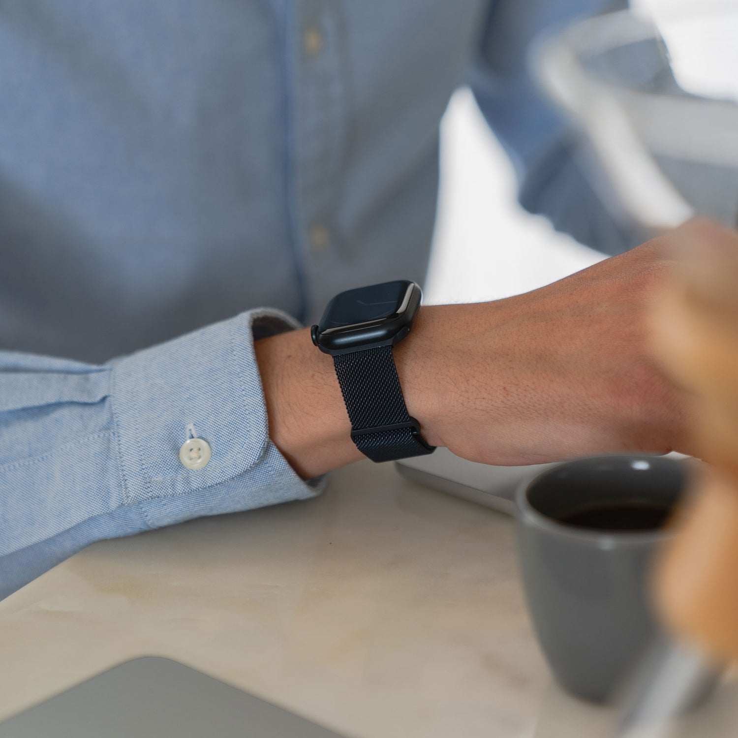 Eine Person trägt ein blaues Langarmhemd und ein Arktisband Apple Watch Milanaise Loop Armband. Sie sitzt mit einer grauen Tasse und einem Laptop an einem Schreibtisch und ihre Hand ruht neben dem Kaffee.
