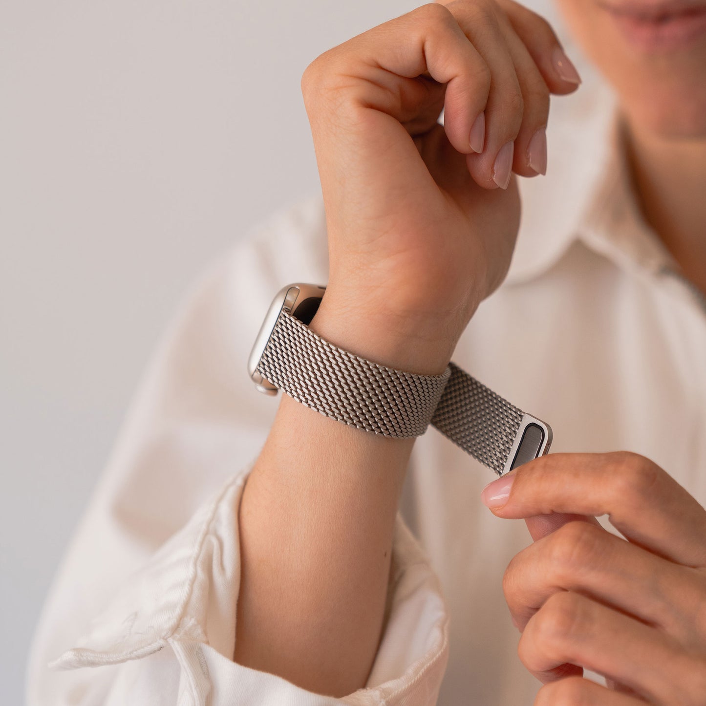 Eine Person passt das arktisband Apple Watch Milanaise Armband „Bold“ in Silber an ihrem Handgelenk an und trägt dabei ein weißes Langarmshirt. Das elegante Edelstahlarmband von arktisband ist mit der Apple Watch kompatibel.