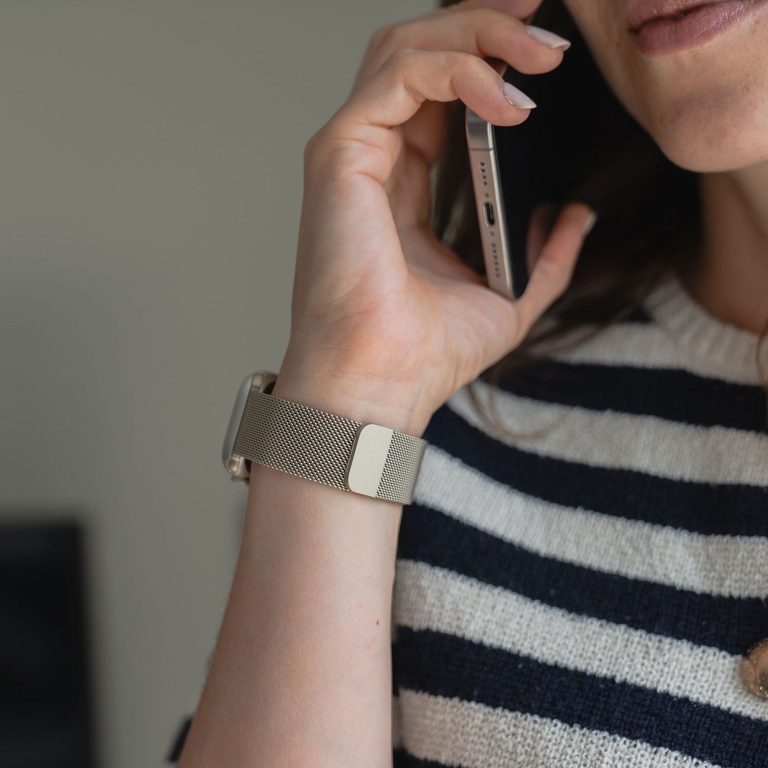 Eine Person, die ein Arktisband Apple Watch Milanaise Loop Armband in Gold und ein schwarz-weiß gestreiftes Oberteil trägt, hält ein Smartphone an ihr Ohr, wobei ihr Gesicht und ihre Hand teilweise sichtbar sind.