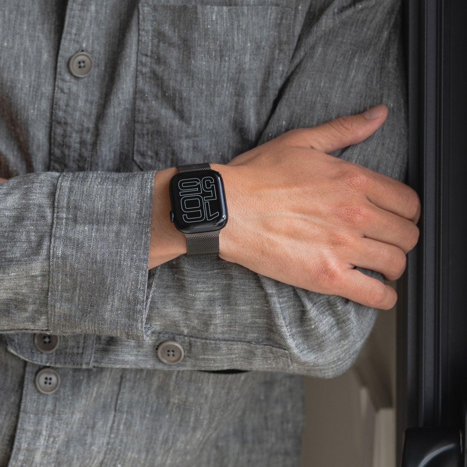 Eine Person in einem grauen Hemd mit Knöpfen verschränkt den rechten Arm und zeigt eine Apple Watch mit einem schwarzen arktisband Apple Watch Milanaise Loop Armband von arktisband und einer großen digitalen Zeitanzeige.