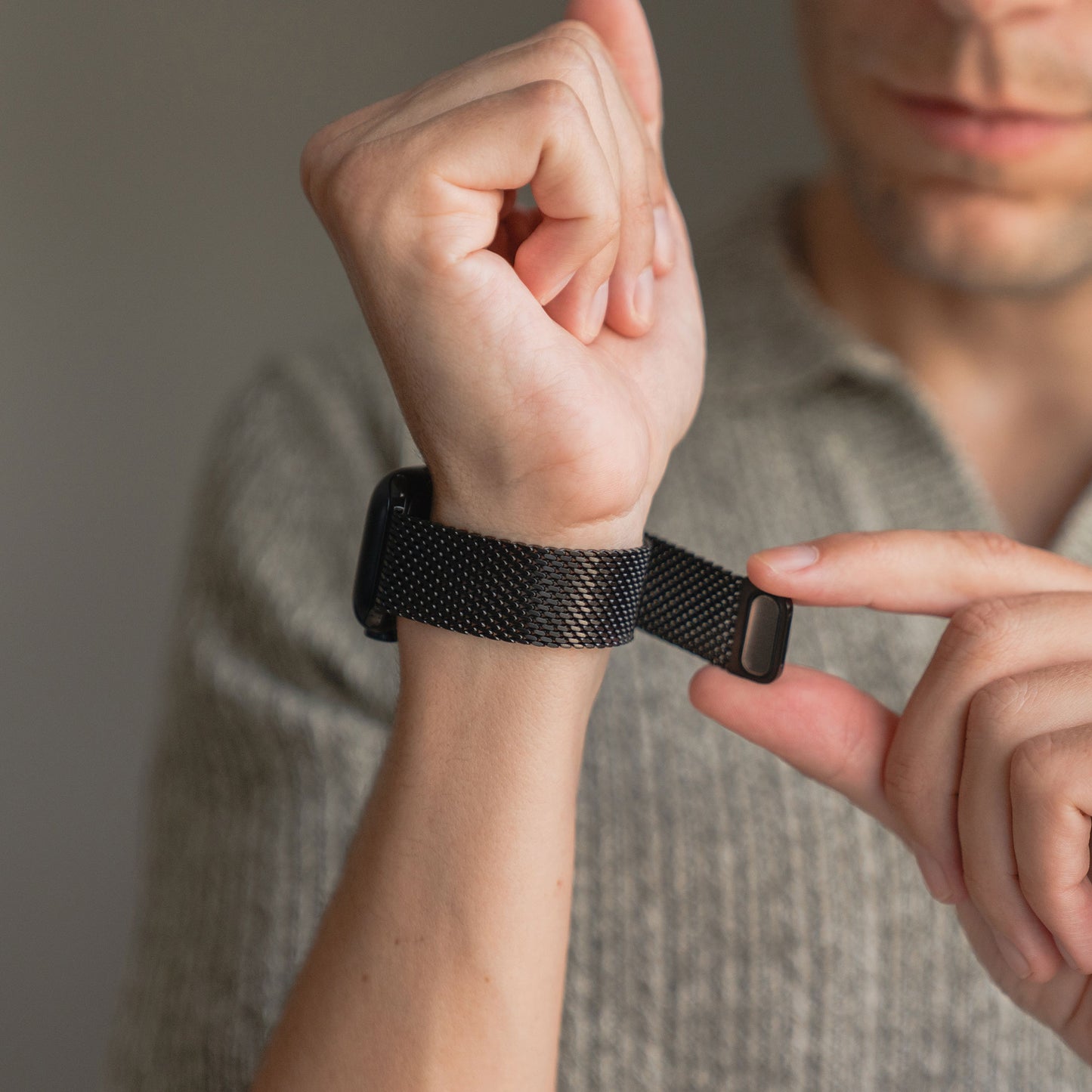 Eine Person befestigt ein arktisband Apple Watch Milanaise Armband "Bold" in Schwarz an ihrer Uhr und trägt ein graues strukturiertes Hemd.