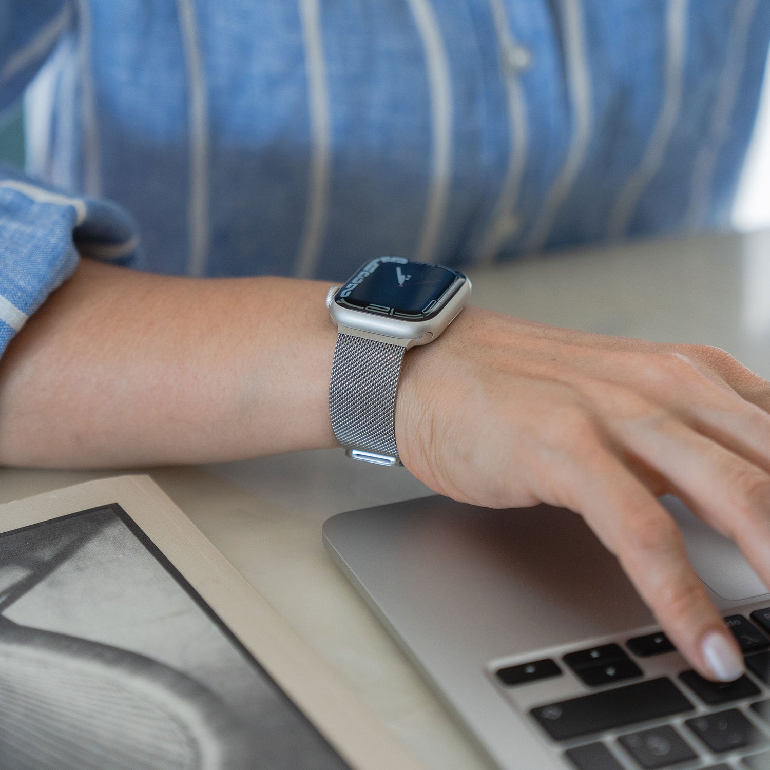 Eine Person trägt ein Arktisband Apple Watch Milanaise Loop Armband in Silber auf einem Laptop, ist mit einem blau-weiß gestreiften Hemd bekleidet und auf dem Tisch ist ein Teil einer offenen Zeitschrift zu sehen.