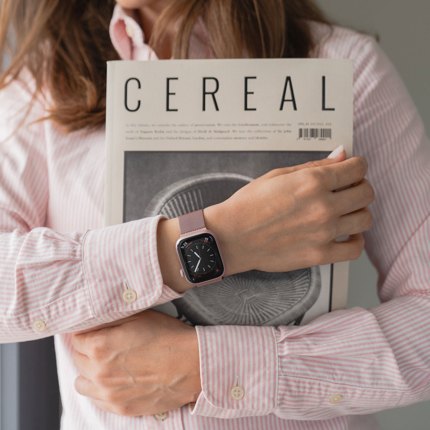 Eine Person in einem rosa gestreiften Hemd trägt ein Arktisband Apple Watch Milanaise Loop Armband und hält ein CEREAL-Magazin mit einem schwarz-weißen Pilz-Cover.