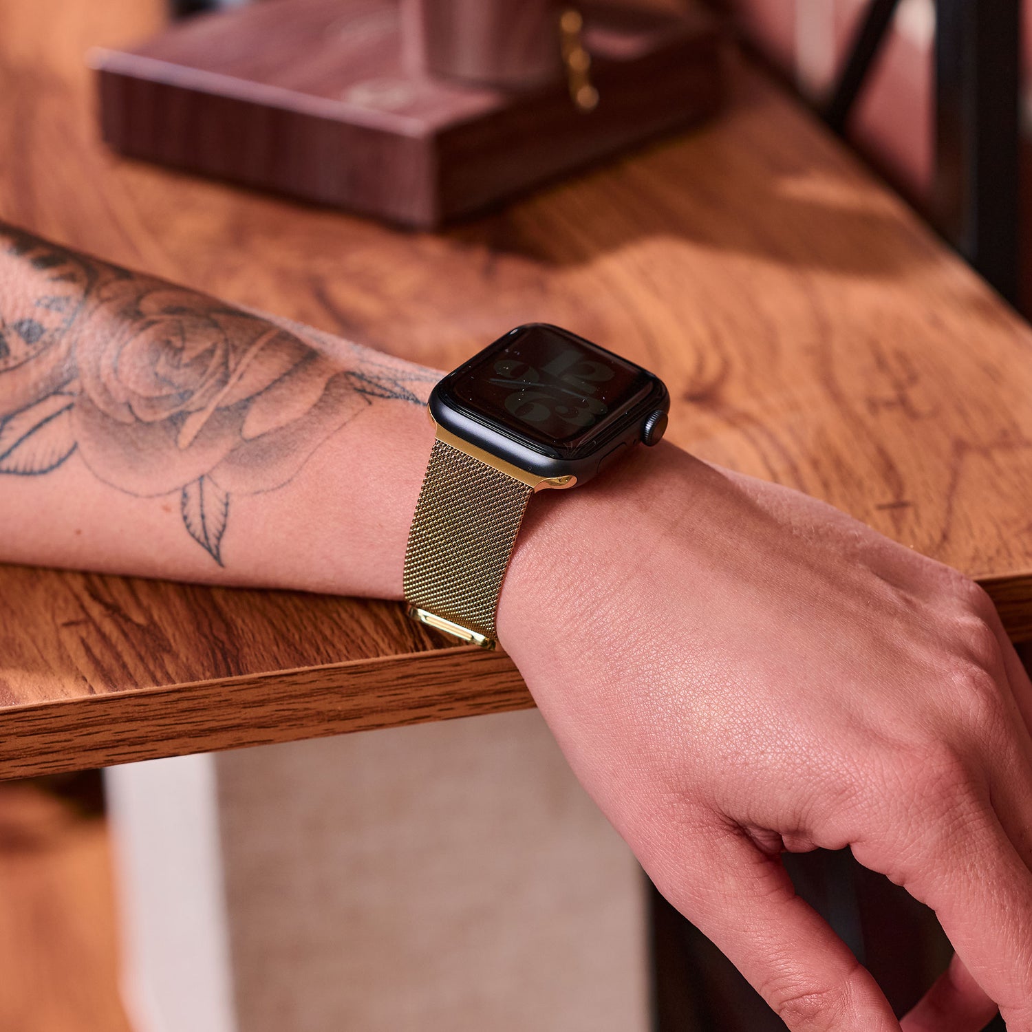 Eine Person mit einem floralen Unterarmtattoo trägt ein Arktisband Apple Watch Milanaise Loop Armband und ruht mit der Hand auf einer Holzoberfläche.
