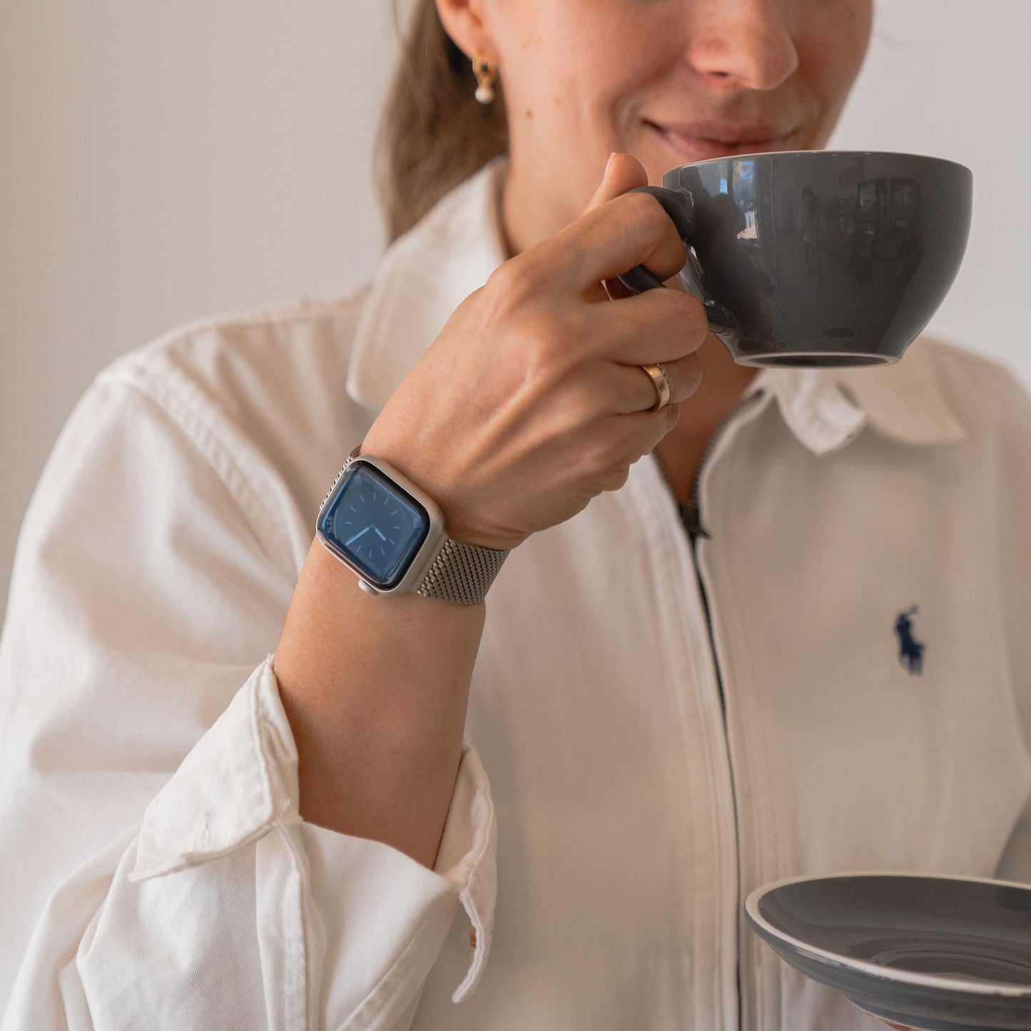 Frau in weißem Hemd hält eine graue Tasse mit Untertasse und trägt das arktisband Apple Watch Milanaise Armband „Bold“ von arktisband am Handgelenk.