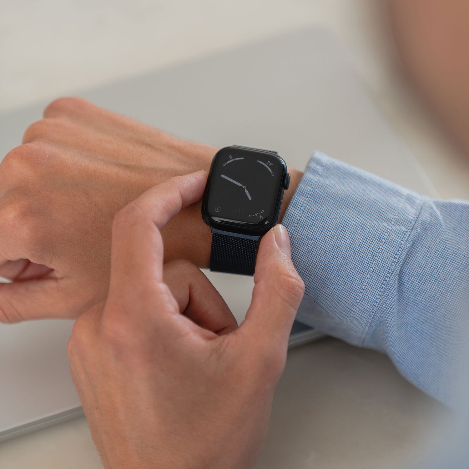 Eine Person in einem blauen Langarmhemd berührt den Bildschirm eines Arktisband Apple Watch Milanaise Loop Armbands an ihrem linken Handgelenk. Auf dem Hintergrund sind ein unscharfer heller Hintergrund und ein Teil eines silbernen Laptops zu sehen.