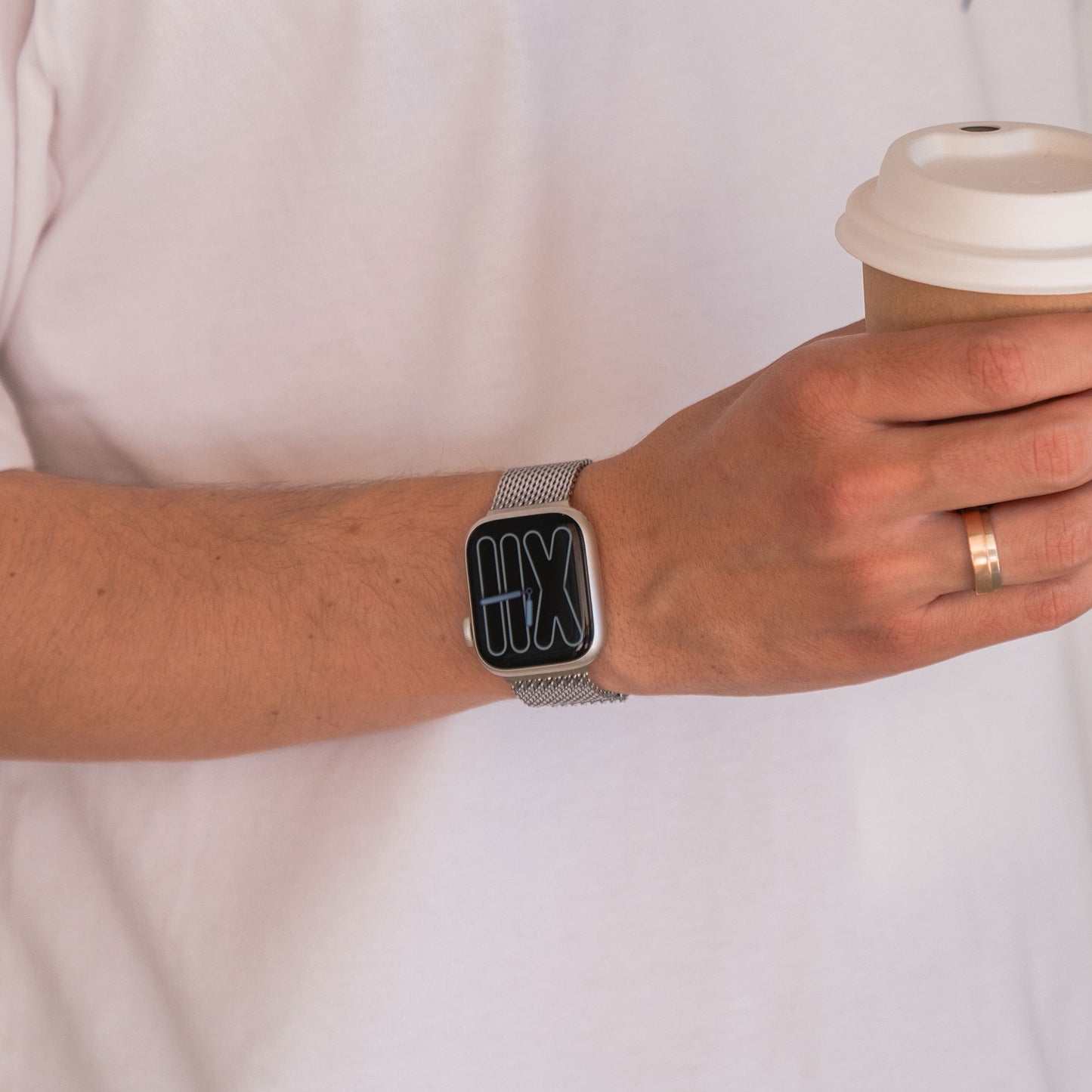 Person, die ein Arktisband Apple Watch Milanaise Armband „Bold“ trägt, eine Kaffeetasse hält und einen goldenen Ring am Finger hat.