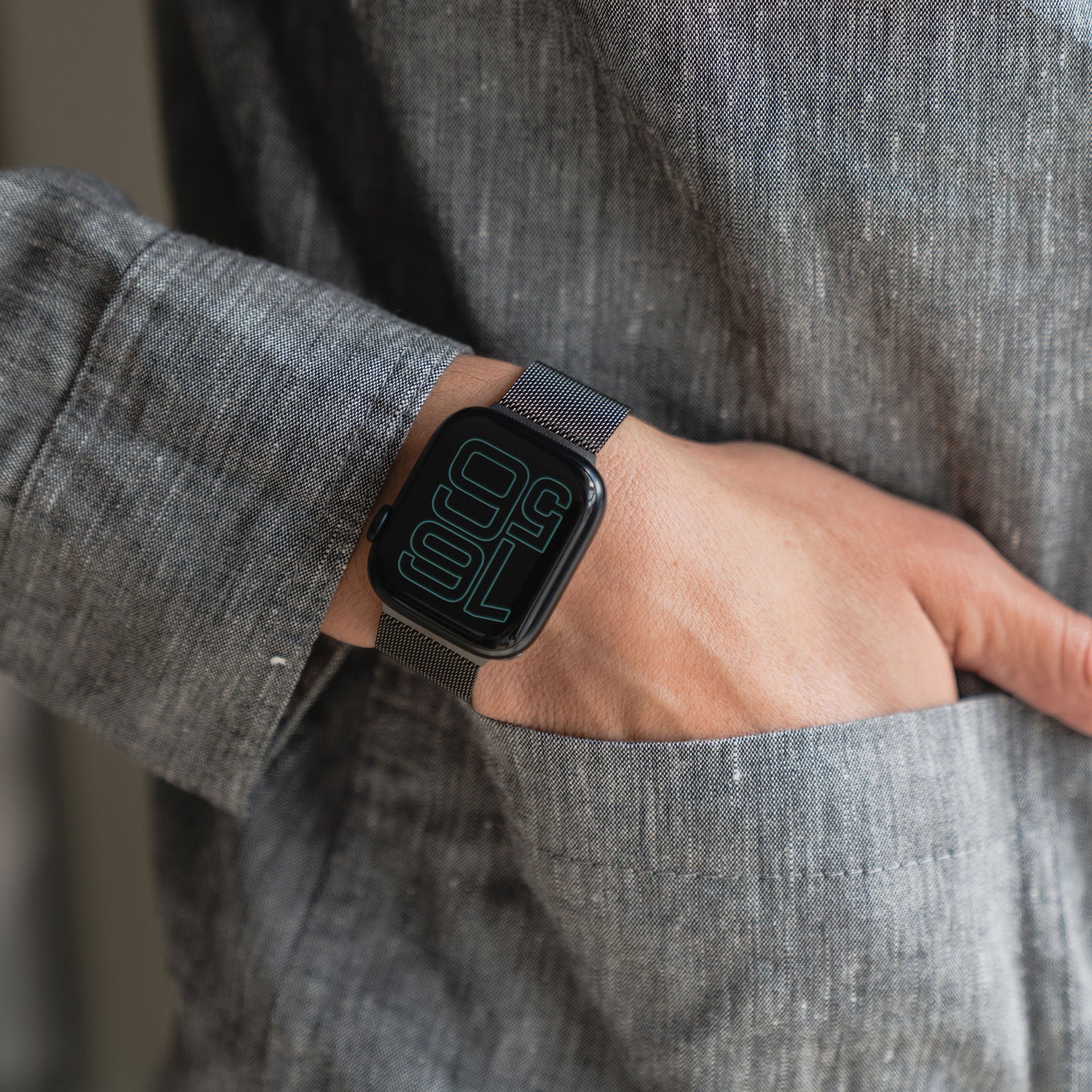 Eine Person in einem grauen Langarmhemd mit einer Hand in der Tasche präsentiert eine Apple Watch mit großer digitaler Zeitanzeige und das elegante Arktisband Apple Watch Milanaise Loop Armband am Handgelenk.