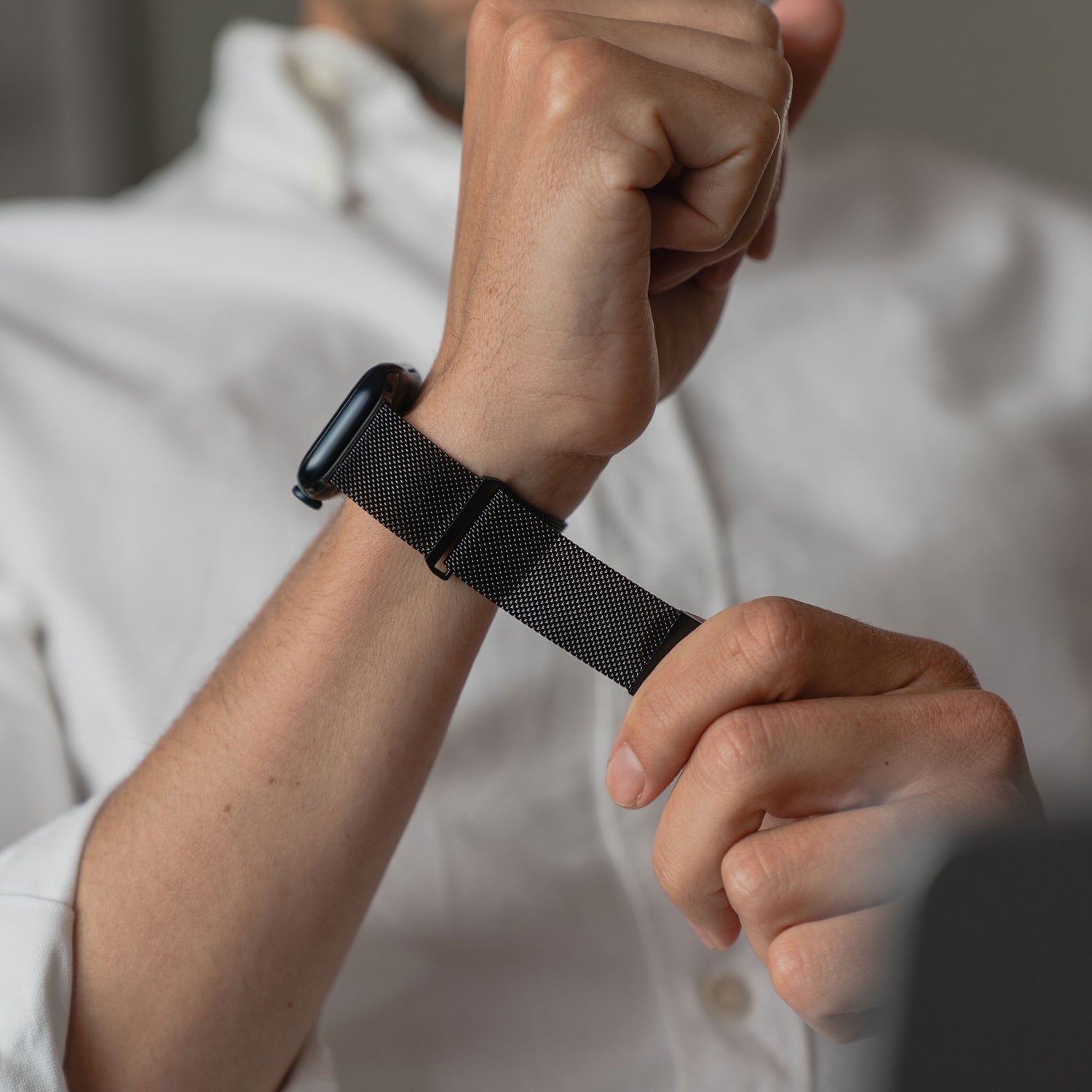 Eine Person in einem weißen Hemd befestigt ein Arktisband Apple Watch Milanaise Loop Armband an ihrer Uhr, wobei der Fokus auf den Zeigern und der Uhr liegt, während ihr Gesicht teilweise außerhalb des Bildes ist.