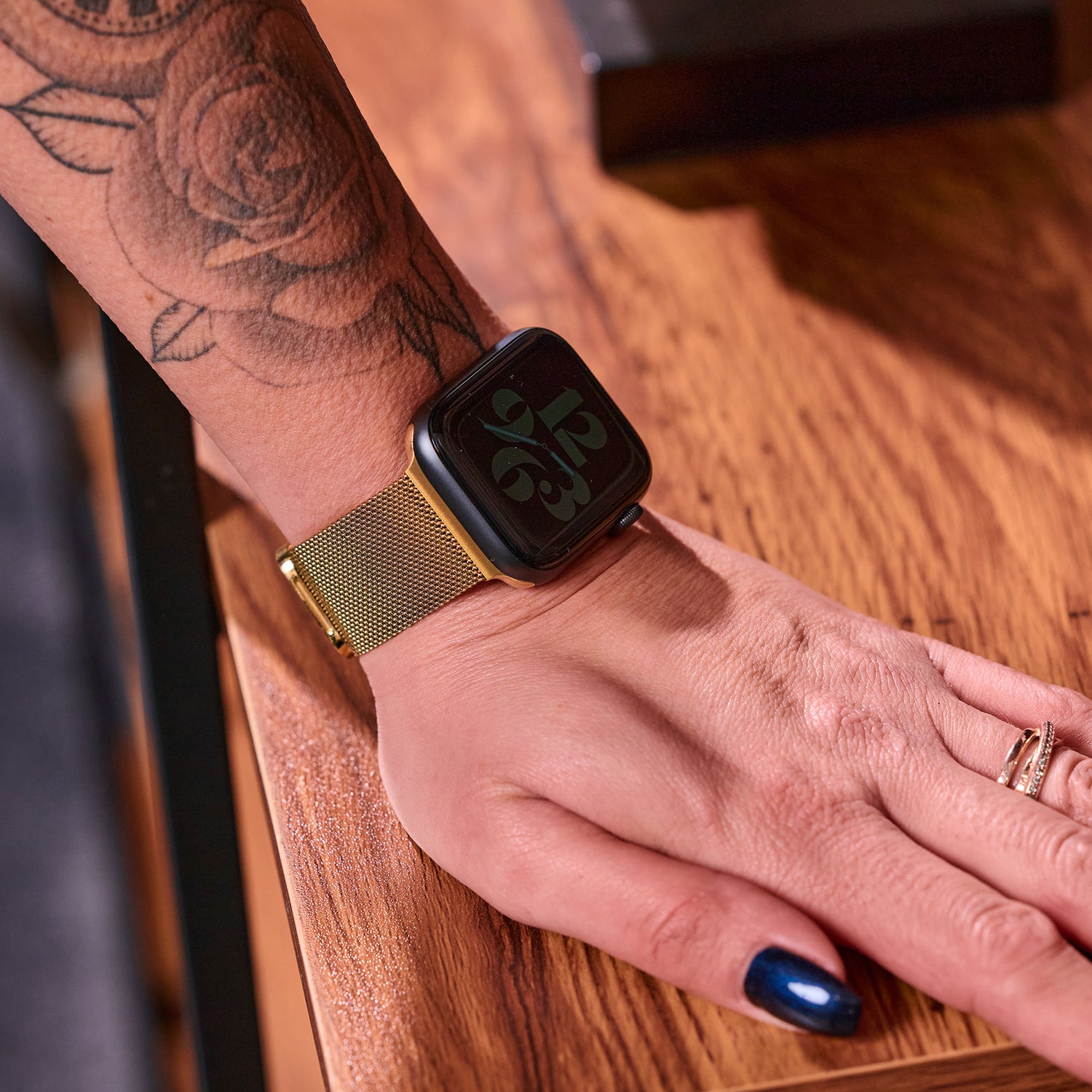 Eine Person mit einem Blumentattoo auf dem Arm trägt ein goldenes Apple Watch Milanaise Loop Armband von Arktisband. Ihre mit einem goldenen Ring verzierte Hand ruht auf einer Holzoberfläche.