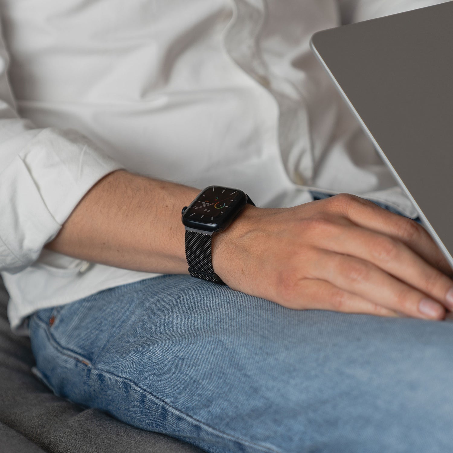 Eine Person in einem weißen Hemd und blauen Jeans sitzt mit einem Laptop da und trägt ein arktisband Apple Watch Milanaise Loop Armband von arktisband am Handgelenk.