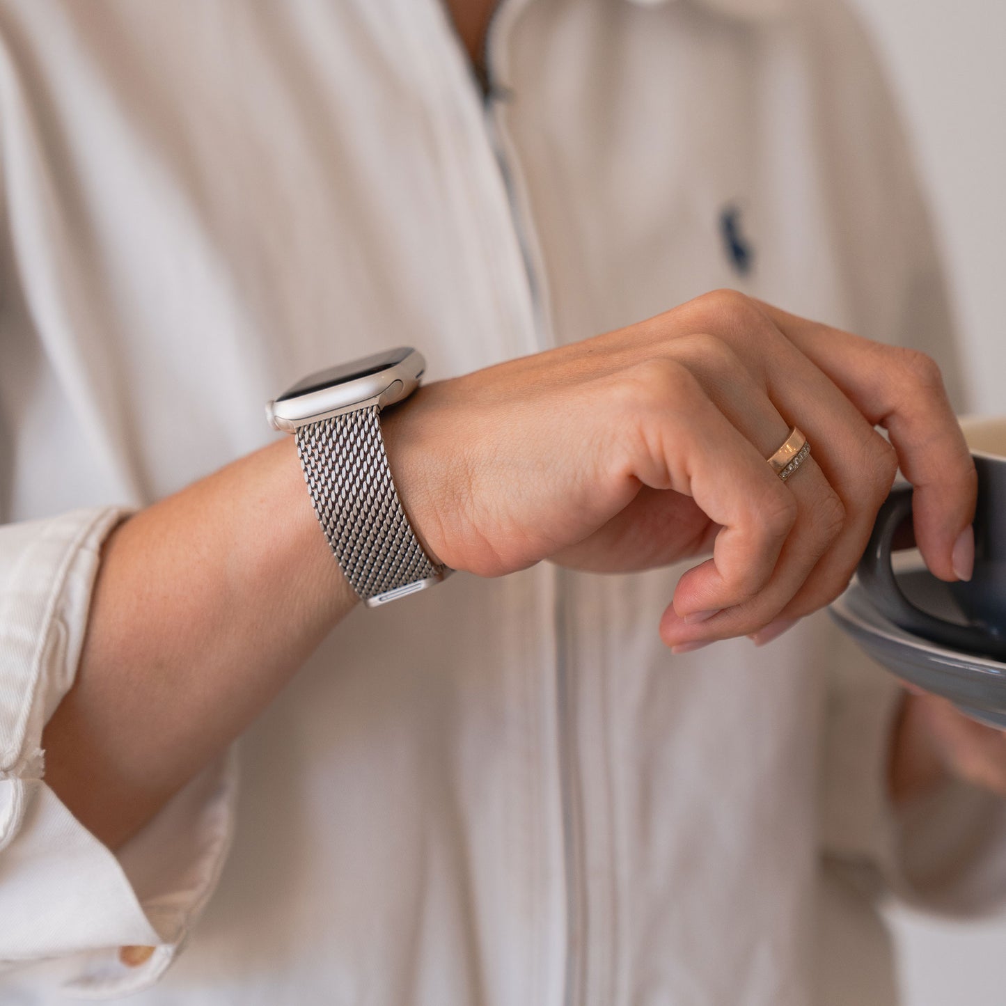 Eine Person trägt ein Arktisband Apple Watch Milanaise Armband „Bold“, hält eine Tasse und trägt ein weißes Hemd mit Reißverschluss.