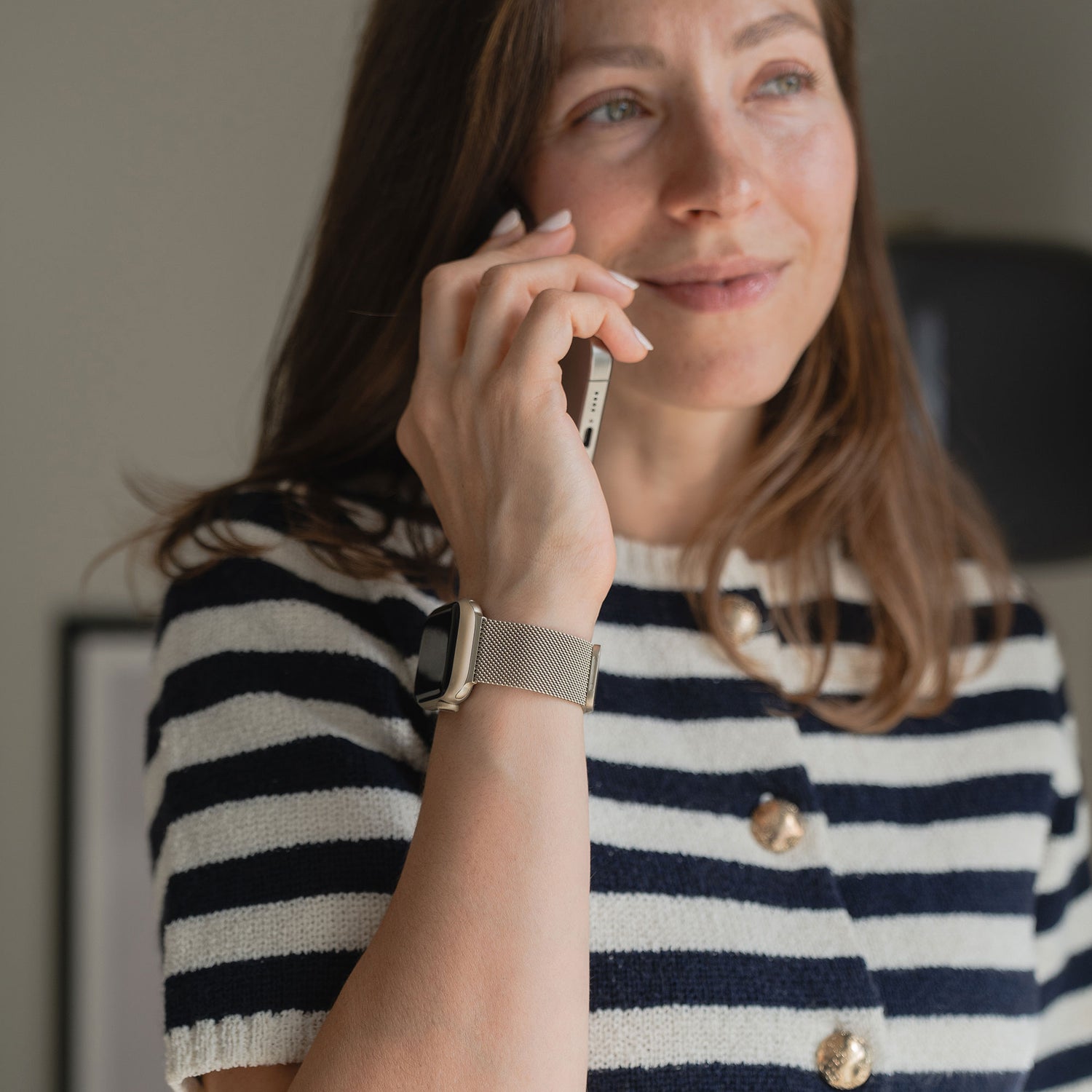 Eine Frau mit langen braunen Haaren, in einem schwarz-weiß gestreiften Hemd und einem Arktisband Apple Watch Milanaise Loop Armband, hält ein Smartphone an ihr Ohr, blickt leicht nach oben und lächelt sanft.