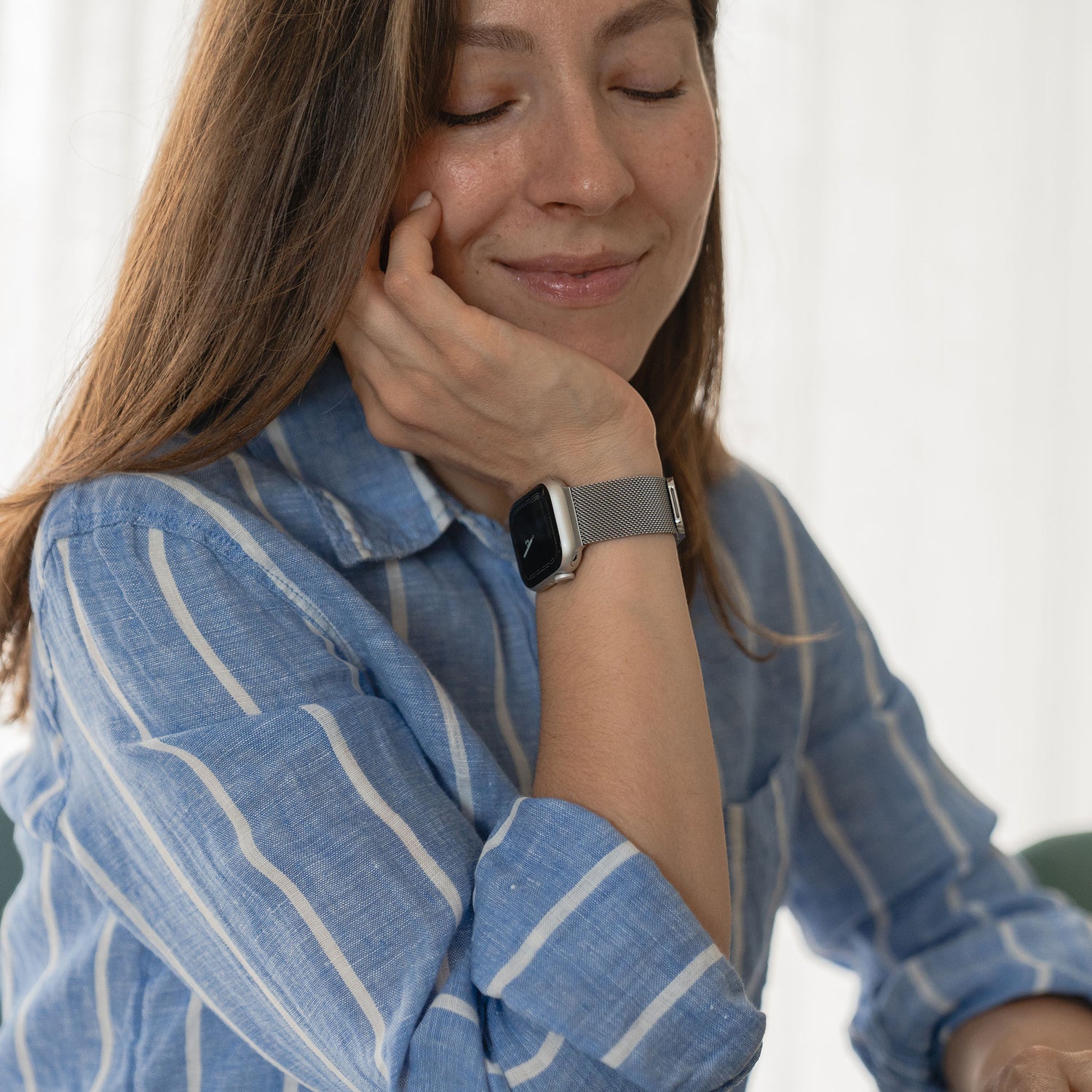 Eine Frau in einem blau-weiß gestreiften Hemd lächelt sanft mit geschlossenen Augen und stützt den Kopf in die Hand. Sie trägt ein arktisband Apple Watch Milanaise Loop Armband von arktisband. Der Hintergrund ist hell und unscharf.