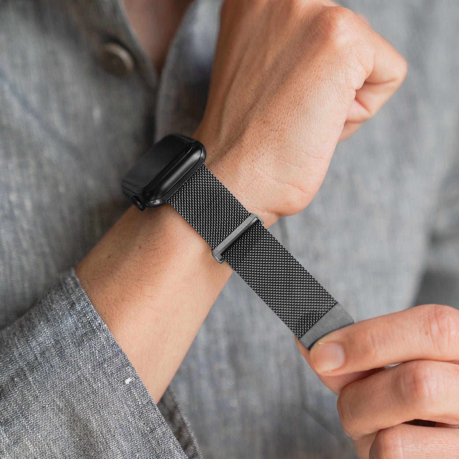 Eine Person in einem grauen Langarmhemd passt das Arktisband Apple Watch Milanaise Loop Armband an ihrer schwarzen Apple Watch an.