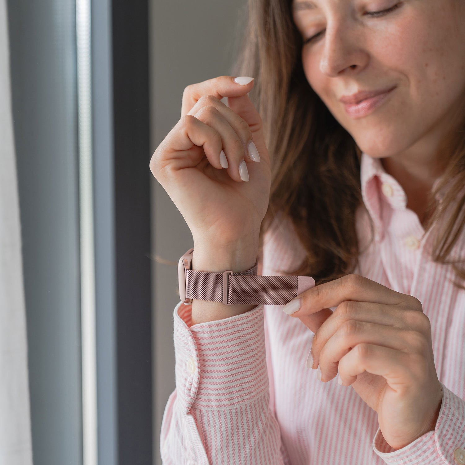 Eine Frau in einem rosa gestreiften Hemd rückt ihre Apple Watch zurecht, die mit einem Arktisband Apple Watch Milanaise Loop Armband ausgestattet ist, während sie in der Nähe eines Fensters steht.