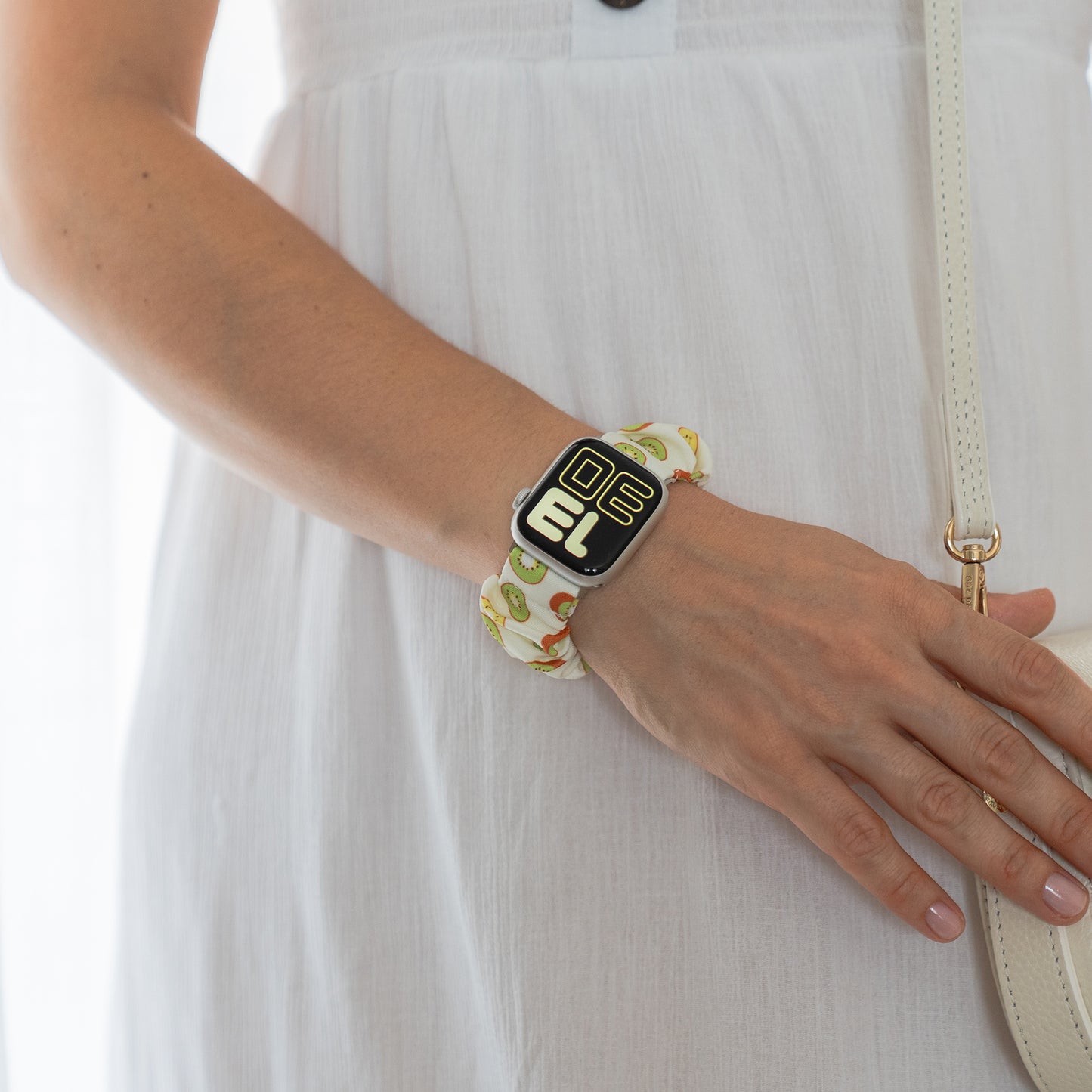 Eine Person in einem weißen Kleid trägt das Arktisband Apple Watch Scrunchie Armband "Elanora" und hält eine cremefarbene Handtasche.