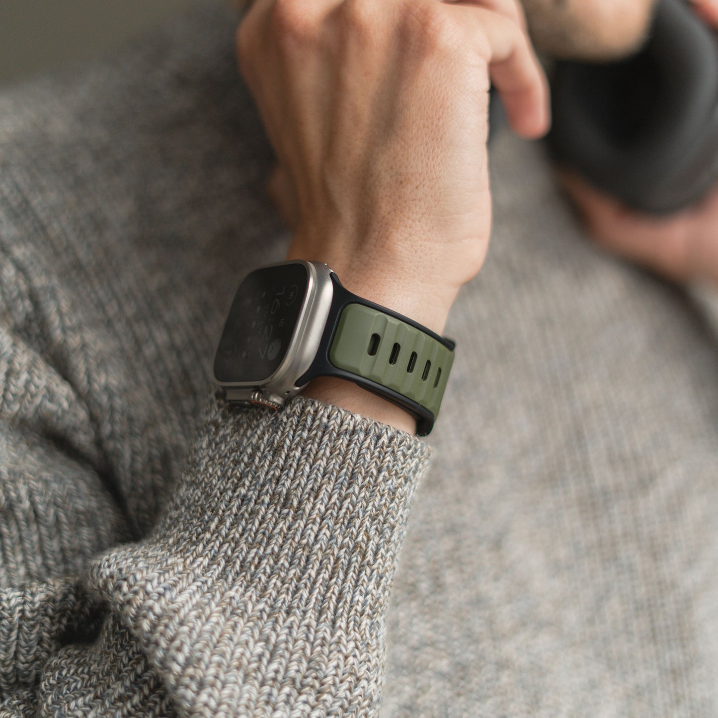 Eine Person in grauem Strickpullover trägt ein arktisband Apple Watch Silikonarmband „Swift“ in Grün-Schwarz, das schweißabsorbierend und atmungsaktiv ist, und hält vermutlich Kopfhörer mit der linken Hand nah ans Gesicht.