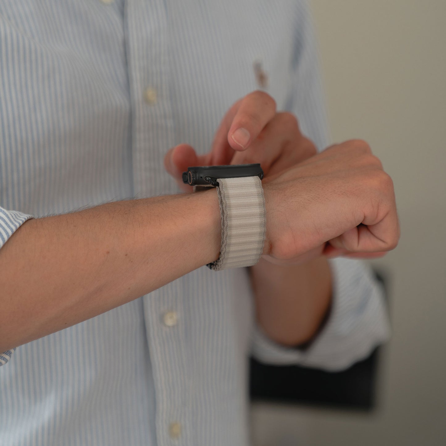 Eine Person in einem hellblau gestreiften Hemd trägt das arktisband Apple Watch Nylonarmband „Venture“ mit geripptem, hellem Band. Der unscharfe Hintergrund passt zu Outdoor-Enthusiasten, die stylische, funktionale Accessoires von arktisband suchen.