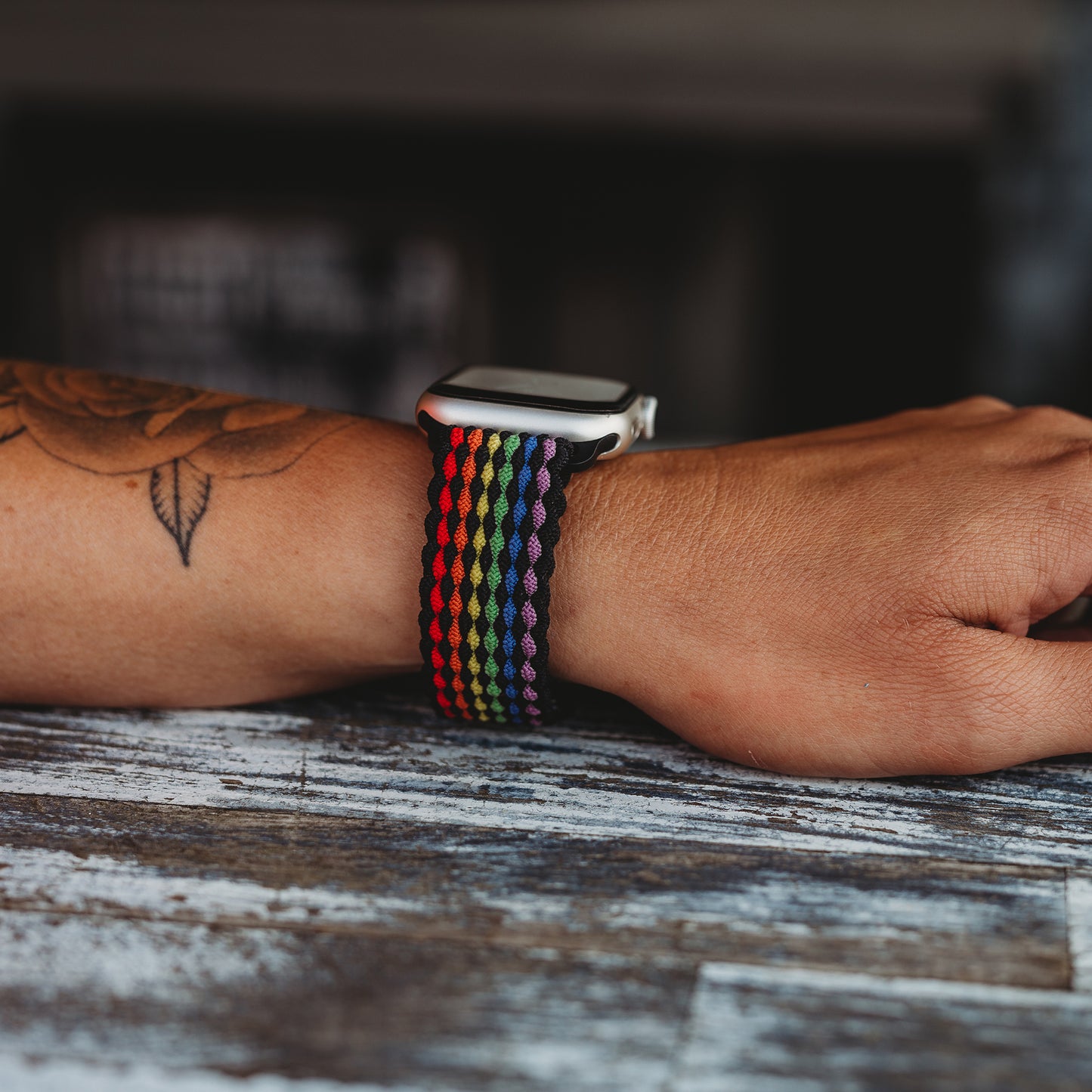 Eine Person mit einem Unterarmtattoo trägt das arktisband Geflochtene Apple Watch Armband „Casual“ Rainbow Edition von arktisband mit Regenbogenstreifen und magnetischem Metallverschluss, während ihr Arm auf einer verwitterten Holzoberfläche ruht.