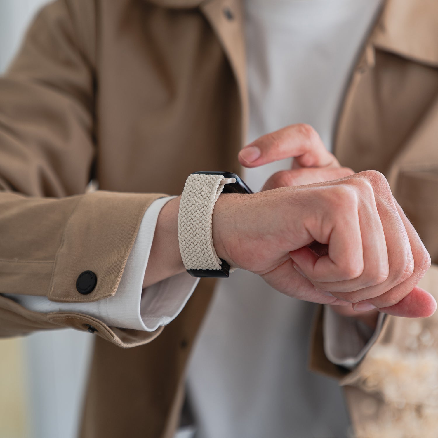 Eine Person in einer hellbraunen Jacke berührt ihr linkes Handgelenk, an dem sich ein geflochtenes Apple Watch Armband „Casual“ mit Magnetverschluss von arktisband mit einem hellen geflochtenen Band befindet; das Bild ist auf ihre Hand und ihr Handgelenk zentriert.