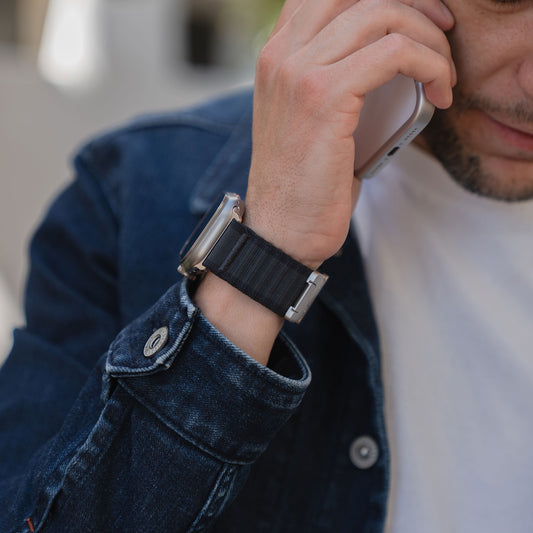 Ein Mann in Jeansjacke telefoniert mit seinem Smartphone und trägt ein arktisband Apple Watch Armband "CoreX" mit einem dunklen Fallschirm-Verschluss-Armband am Handgelenk.