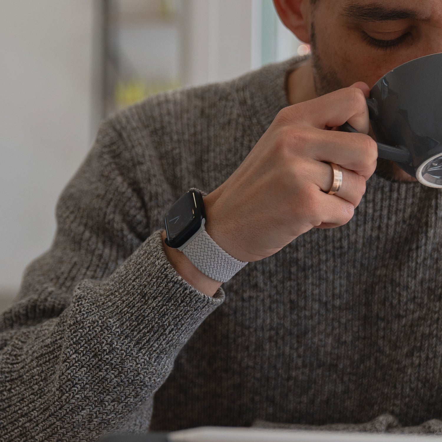 Ein Mann trägt ein geflochtenes Apple Watch Armband „Casual“ mit Magnetverschluss von Arktisband und einen Ehering und nippt in einem grauen Strickpullover an einer Tasse.