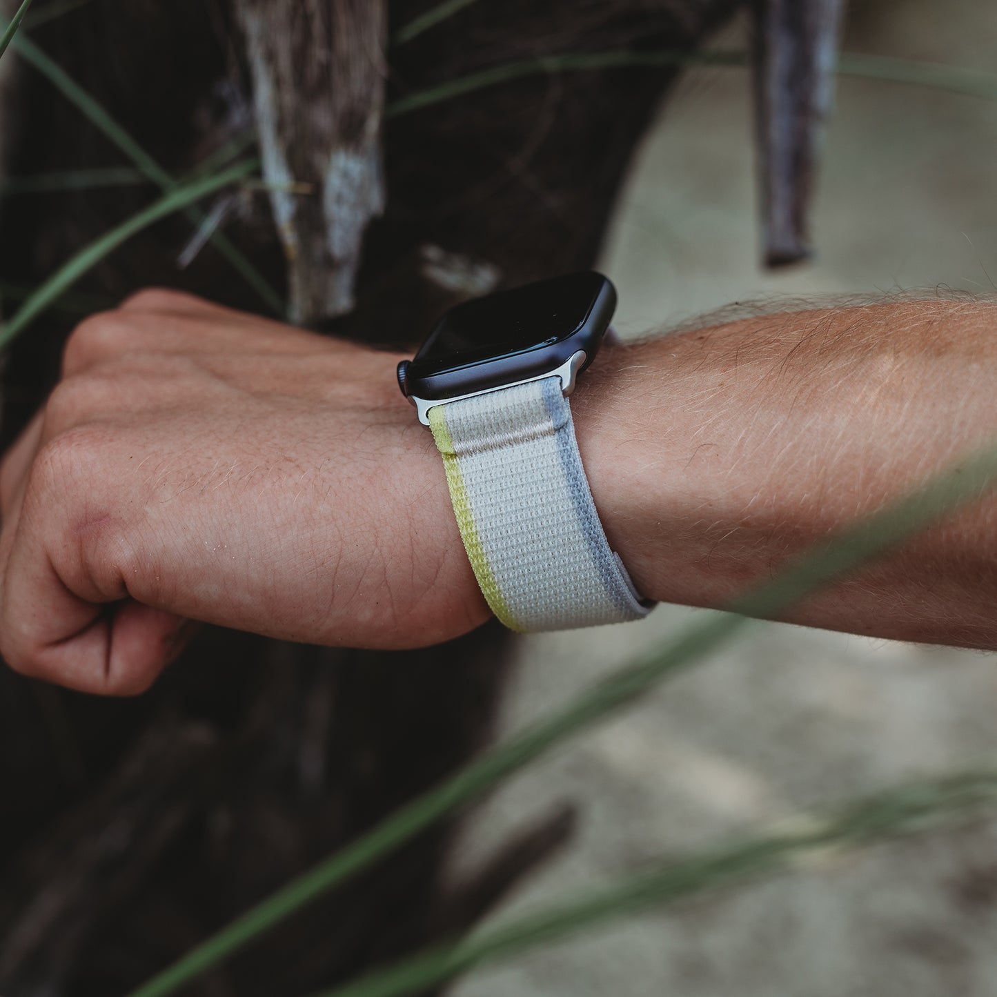 Das Handgelenk einer Person, die das arktisband Apple Watch Armband „Trail“ von arktisband aus weichem Polyester mit Klettverschluss trägt, befindet sich im Freien in der Nähe eines Baums und grünen Grases.