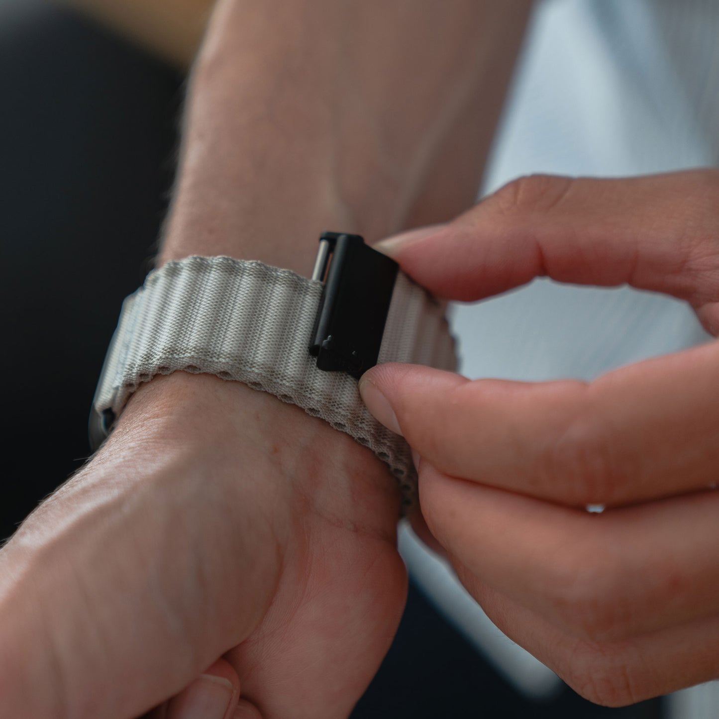 Eine Nahaufnahme zeigt jemanden, der das graue Arktisband Apple Watch Nylonarmband „Venture“ mit schwarzem Verschluss an seinem Handgelenk befestigt. Der unscharfe Hintergrund hebt das stylische Armband hervor – ideal für Outdoor-Enthusiasten.