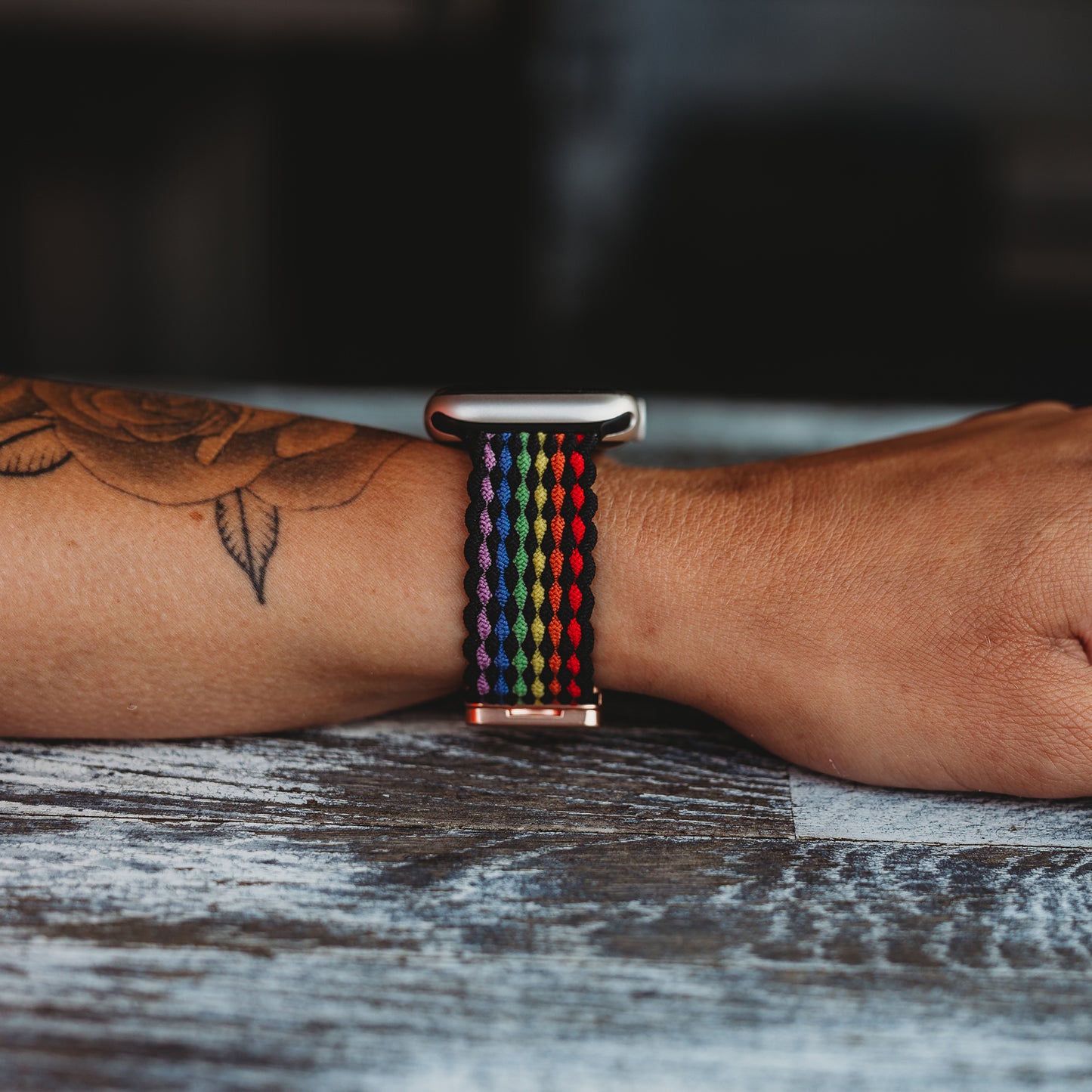 Eine Person mit einem floralen Unterarmtattoo trägt das arktisband Geflochtene Apple Watch Armband „Casual“ Rainbow Edition mit roten, schwarzen, blauen, violetten und grünen Streifen und einem magnetischen Metallverschluss, der auf einer Holzoberfläche ruht.