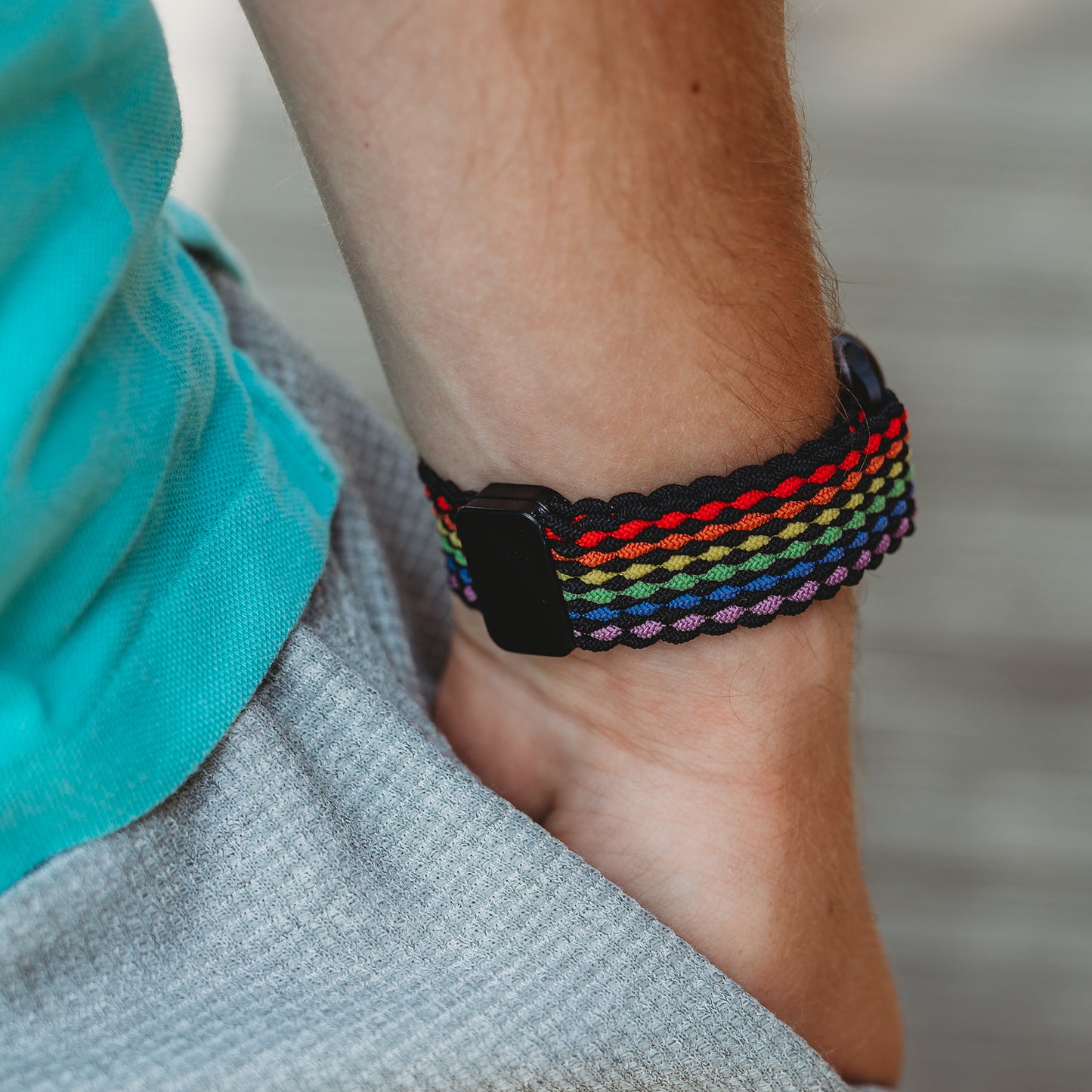 Eine Person, die ein geflochtenes Apple Watch Armband „Casual“ Rainbow Edition mit magnetischem Metallverschluss und ein blaugrünes Hemd trägt, steht mit der Hand in der Tasche einer grauen Hose.