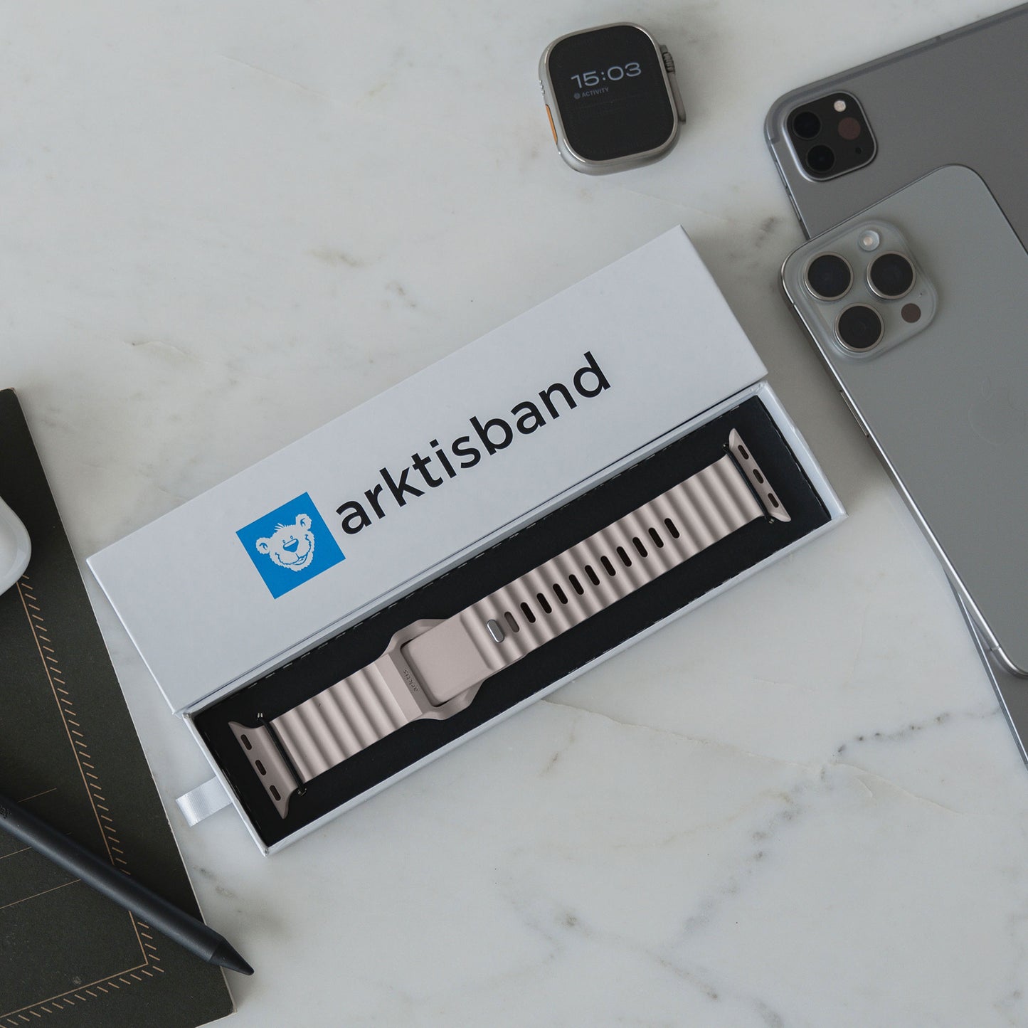 Ein verpacktes Arktisband Apple Watch Sportarmband „Motion“ wird auf einer weißen Marmoroberfläche neben einer Smartwatch, einem silbernen Smartphone, einem Stift auf einem Notizbuch und einem Tablet präsentiert.