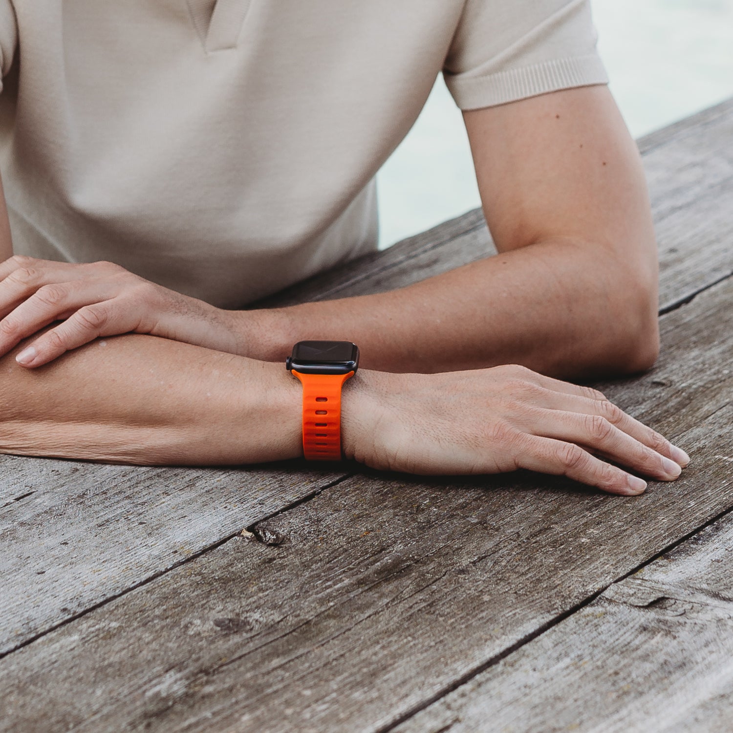Eine Person in einem kurzärmeligen beigen Hemd ruht mit den Armen auf einem verwitterten Holztisch und zeigt an ihrem Handgelenk ein arktisband Apple Watch Silikonarmband „Active“ von arktisband.