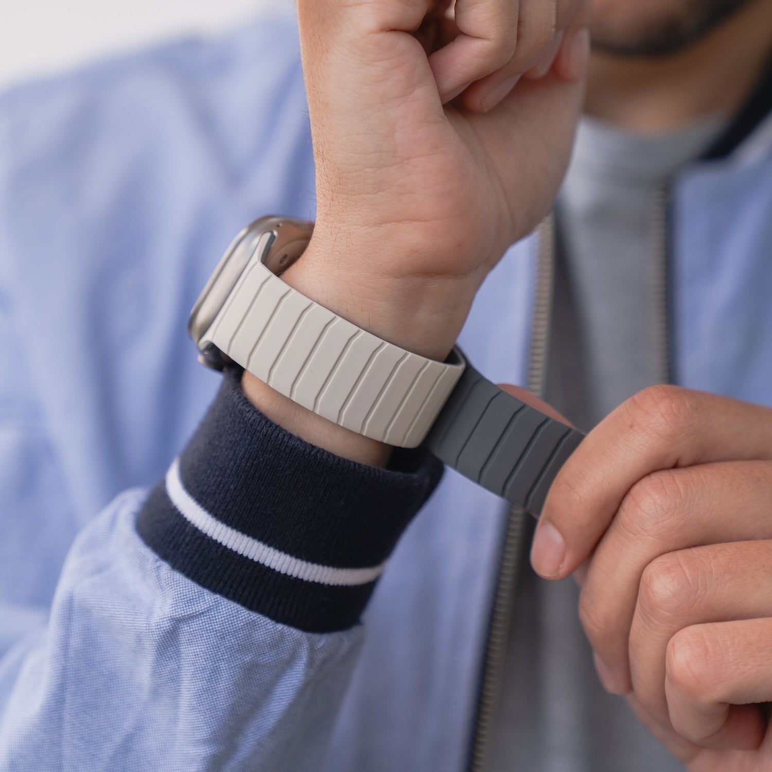 Eine Person befestigt das Arktisband Apple Watch Magnetarmband „MagFlow“ mit beigem Silikonband am Handgelenk und trägt dabei eine hellblaue Jacke.