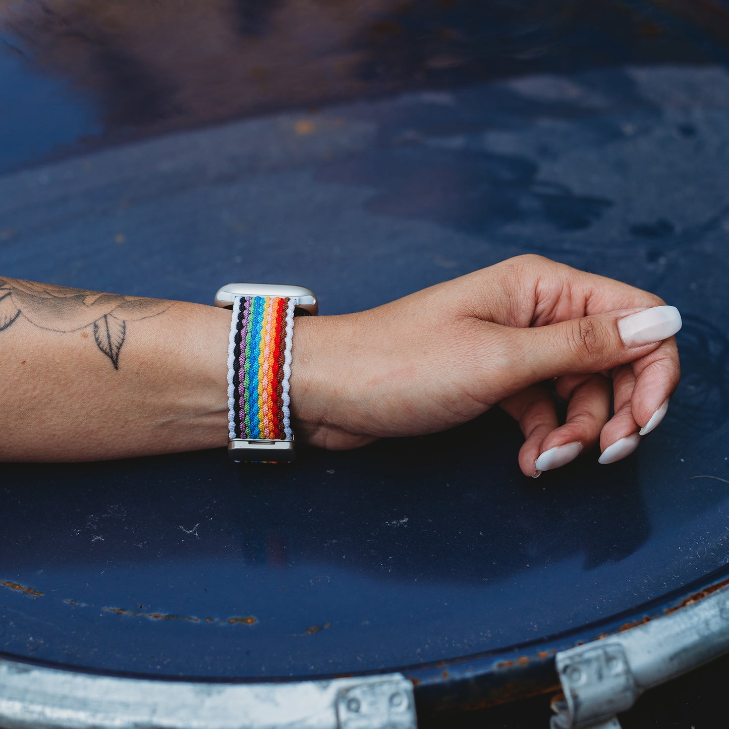Eine Hand mit langen, manikürten Nägeln und einem sichtbaren Tattoo ruht auf blauem Metall und trägt das arktisband Geflochtene Apple Watch Armband "Casual" Rainbow Edition mit magnetischem Metallverschluss.