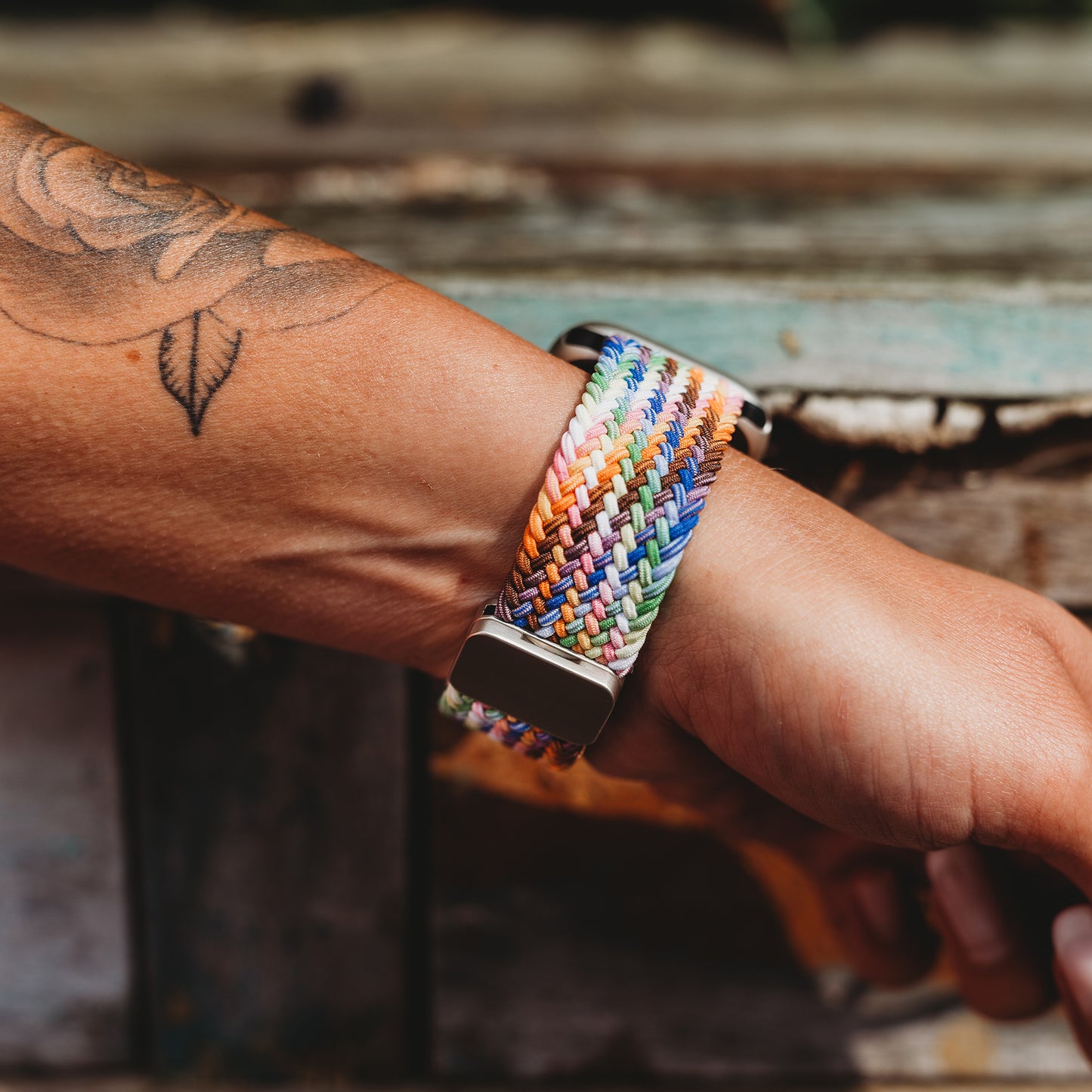 Eine Nahaufnahme eines Handgelenks mit einem schwarzen Blatttattoo, das das von Arktisband geflochtene Apple Watch Armband „Casual“ Rainbow Edition mit magnetischem Metallverschluss trägt, vor verwittertem Holz.