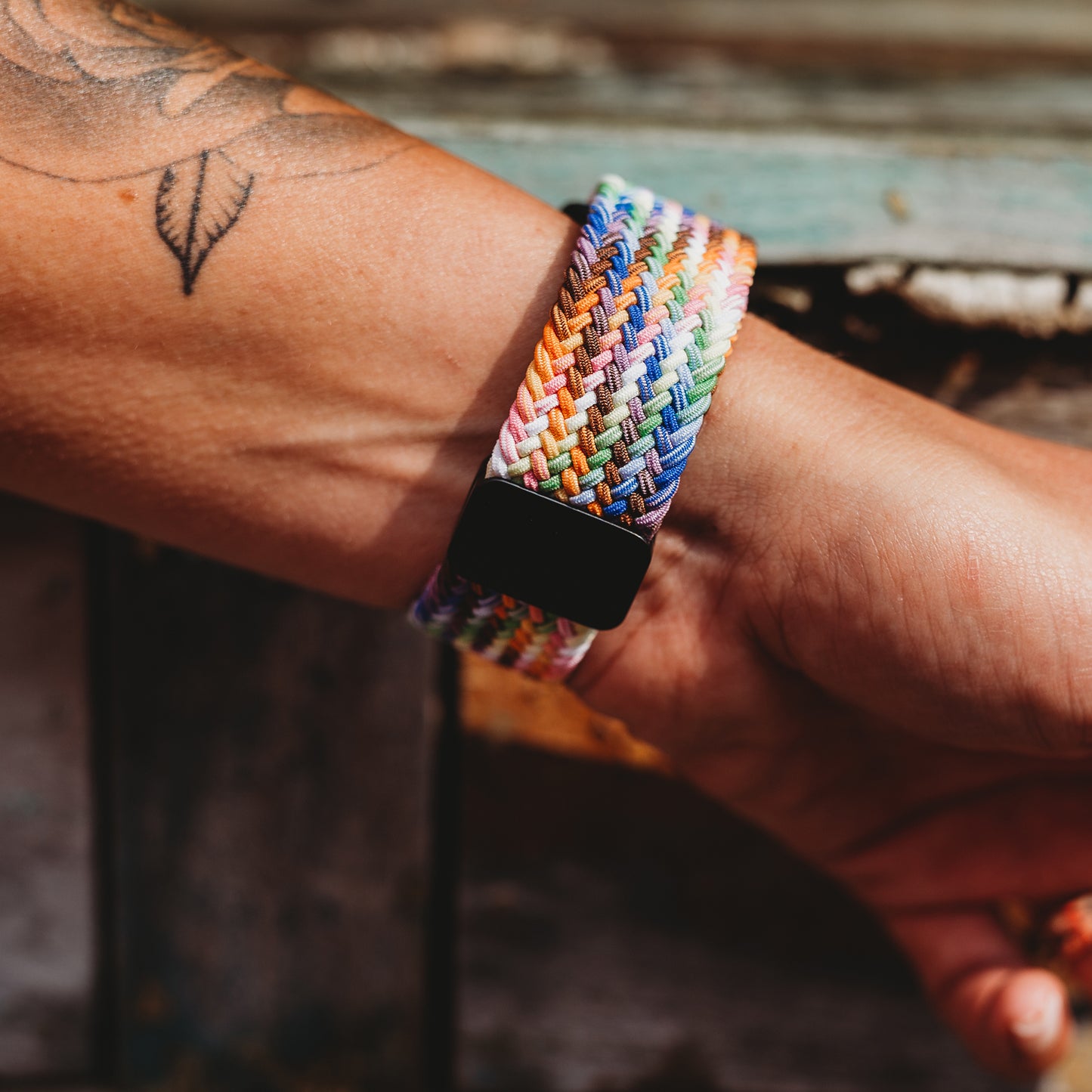 Jemand trägt ein arktisband Geflochtenes Apple Watch Armband „Casual“ Rainbow Edition mit schwarzem Magnetverschluss. Sein Arm mit Blatttattoo ruht auf verwittertem Holz.