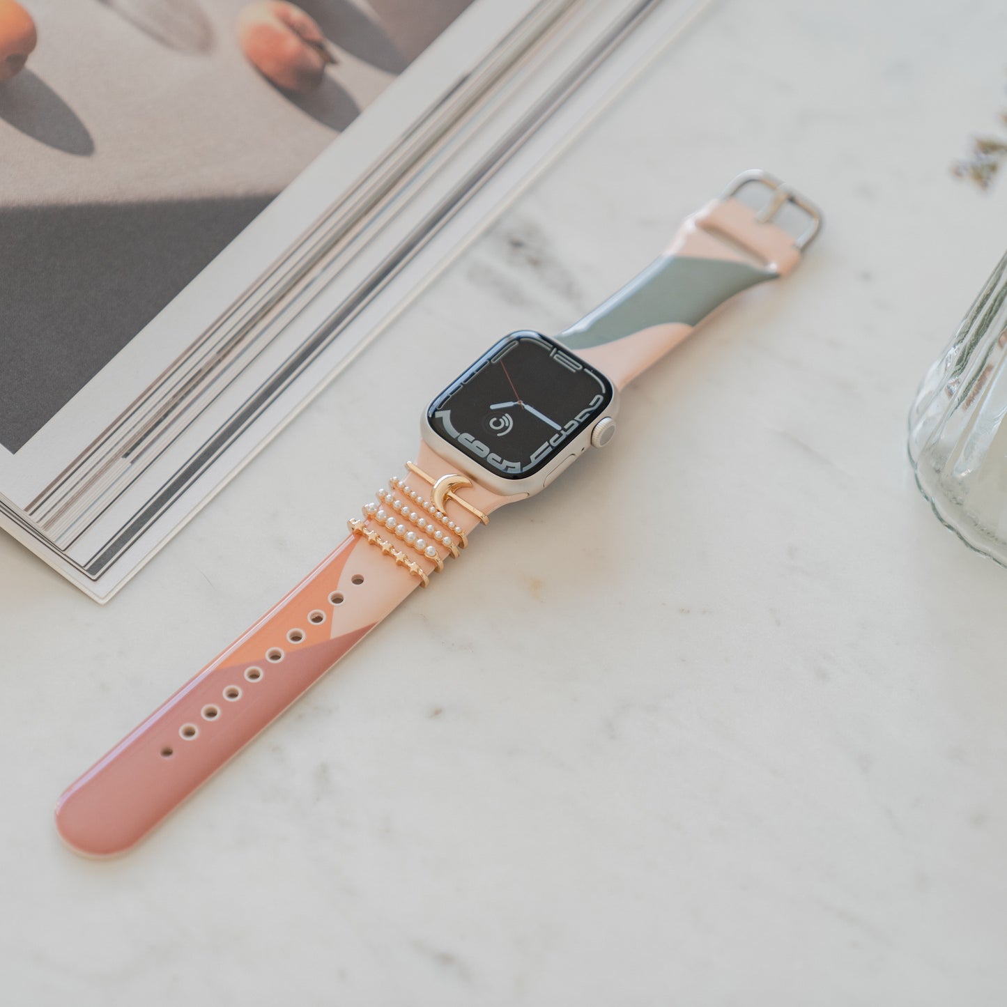 Die arktisband Apple Watch Charms „Moon“ in Gold von arktisband sind auf einer Smartwatch mit pfirsich- und grauem Armband abgebildet, die auf einer weißen Marmoroberfläche neben einer Zeitschrift und einer Glasvase liegt.