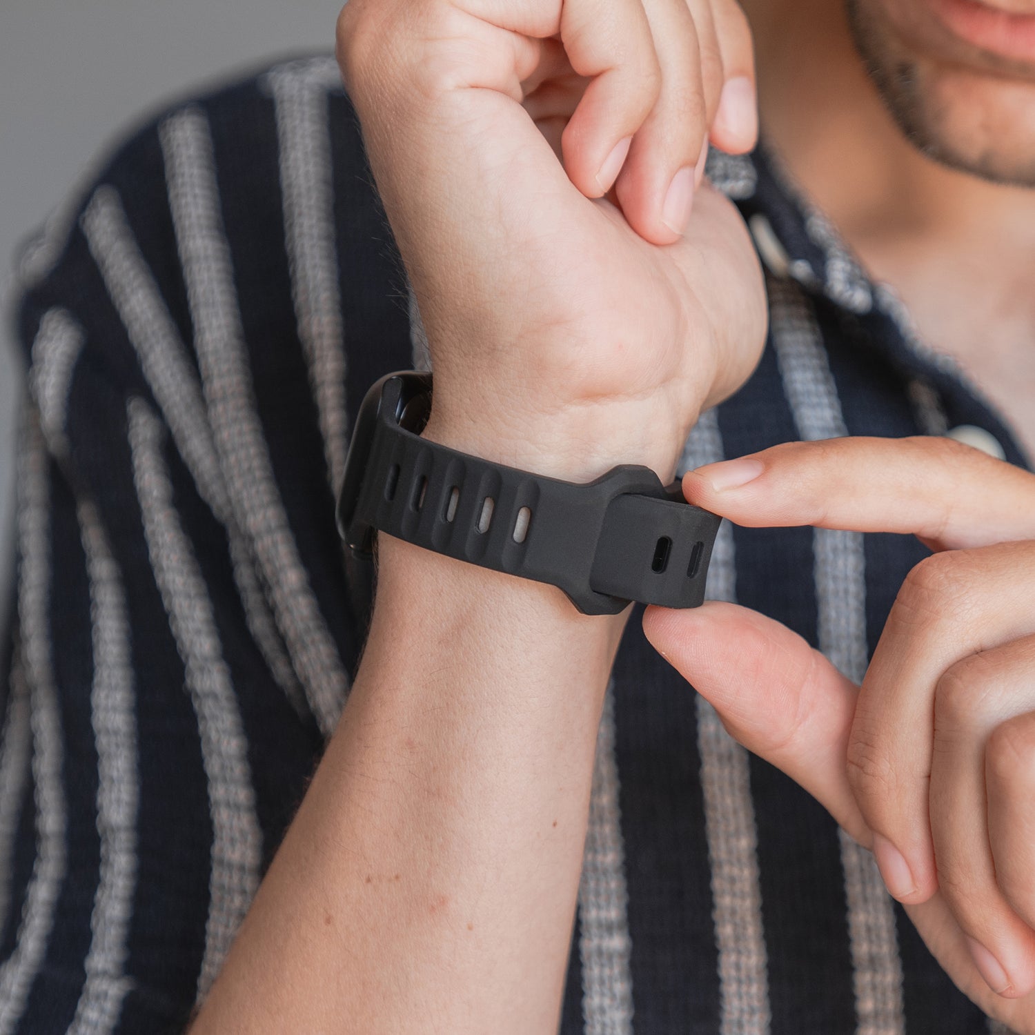 Eine Person mit einem schwarz-weiß gestreiften Hemd befestigt mit der rechten Hand ein arktisband Apple Watch Silikonarmband „Active“ von arktisband mit atmungsaktivem Design am linken Handgelenk.