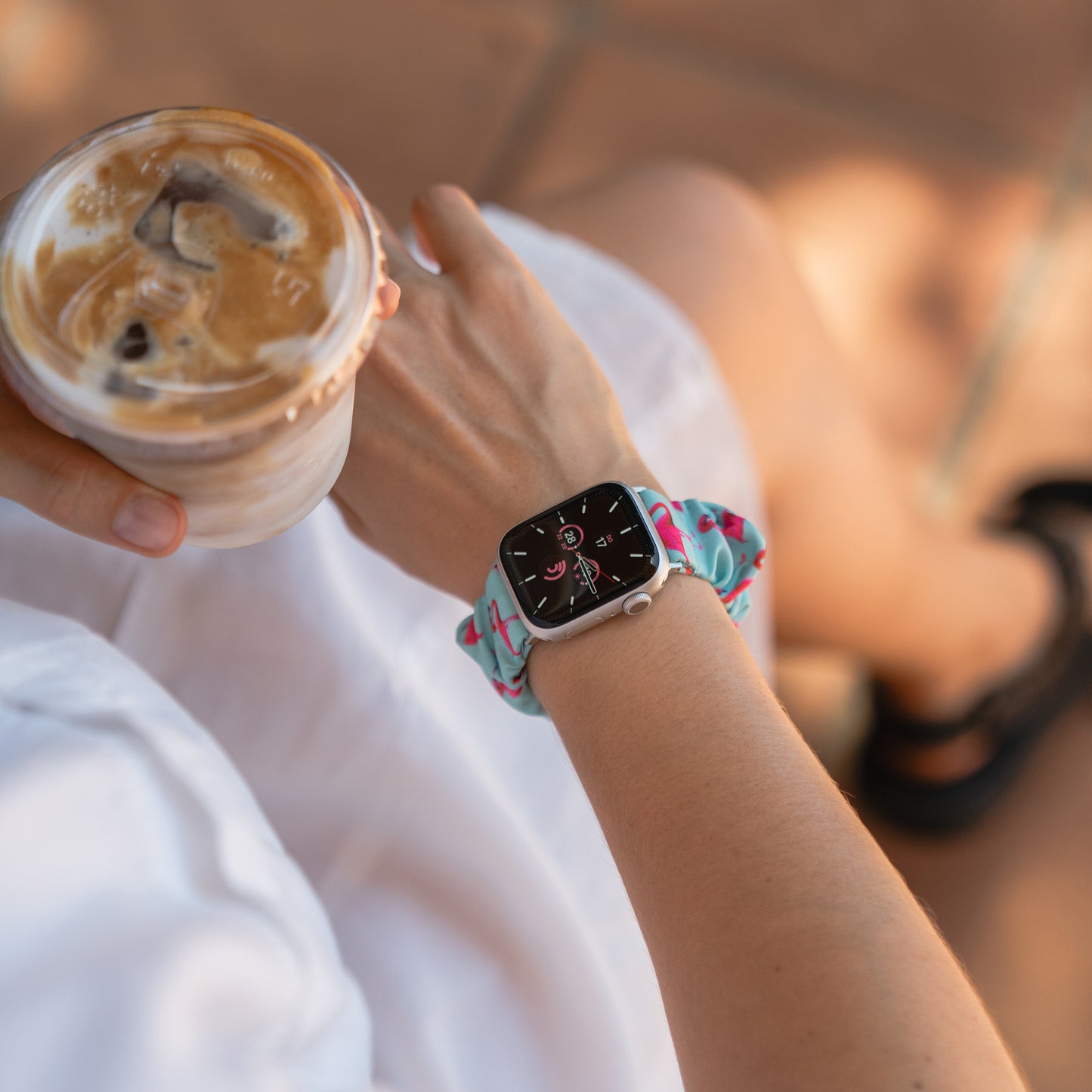 Person mit Eiskaffee in der Hand, die ein arktisband Apple Watch Scrunchie Armband "Elanora" mit einem bunten Armband trägt und im Sonnenlicht sitzt.