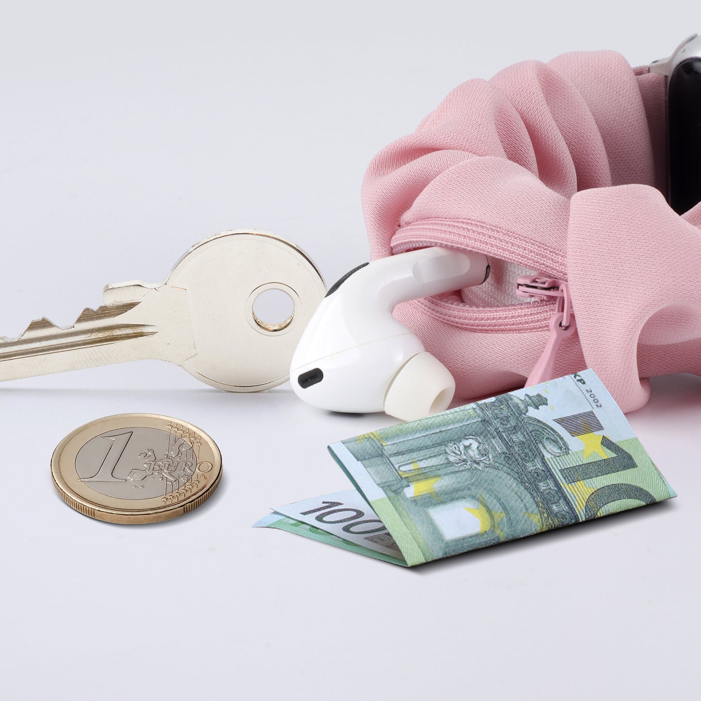 Ein Schlüssel, eine Euro-Münze, ein 100-Euro-Schein und ein Ohrstöpsel befinden sich neben einer rosafarbenen Reißverschlusstasche und dem arktisband Apple Watch Scrunchie Armband "Elanora".